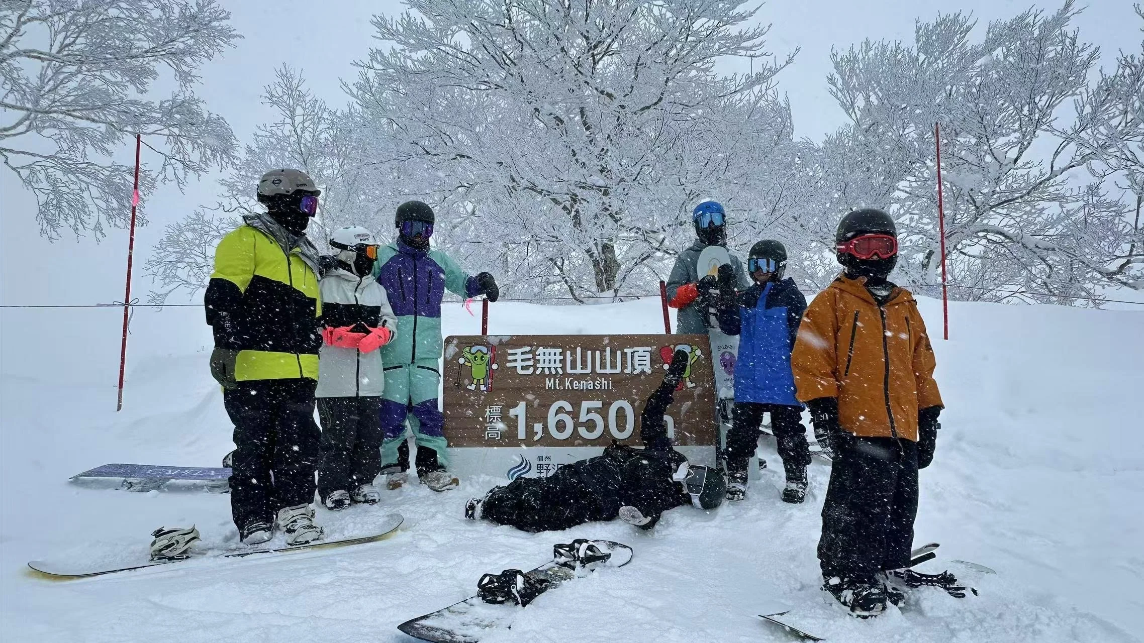 A group of eight skiers and snowboarders in colorful winter gear posing for a photo at the summit of Mt. Kenashi, which is 1,650 meters high. They are standing behind a brown sign with white text and cartoon characters, surrounded by snow-covered trees and falling snow.