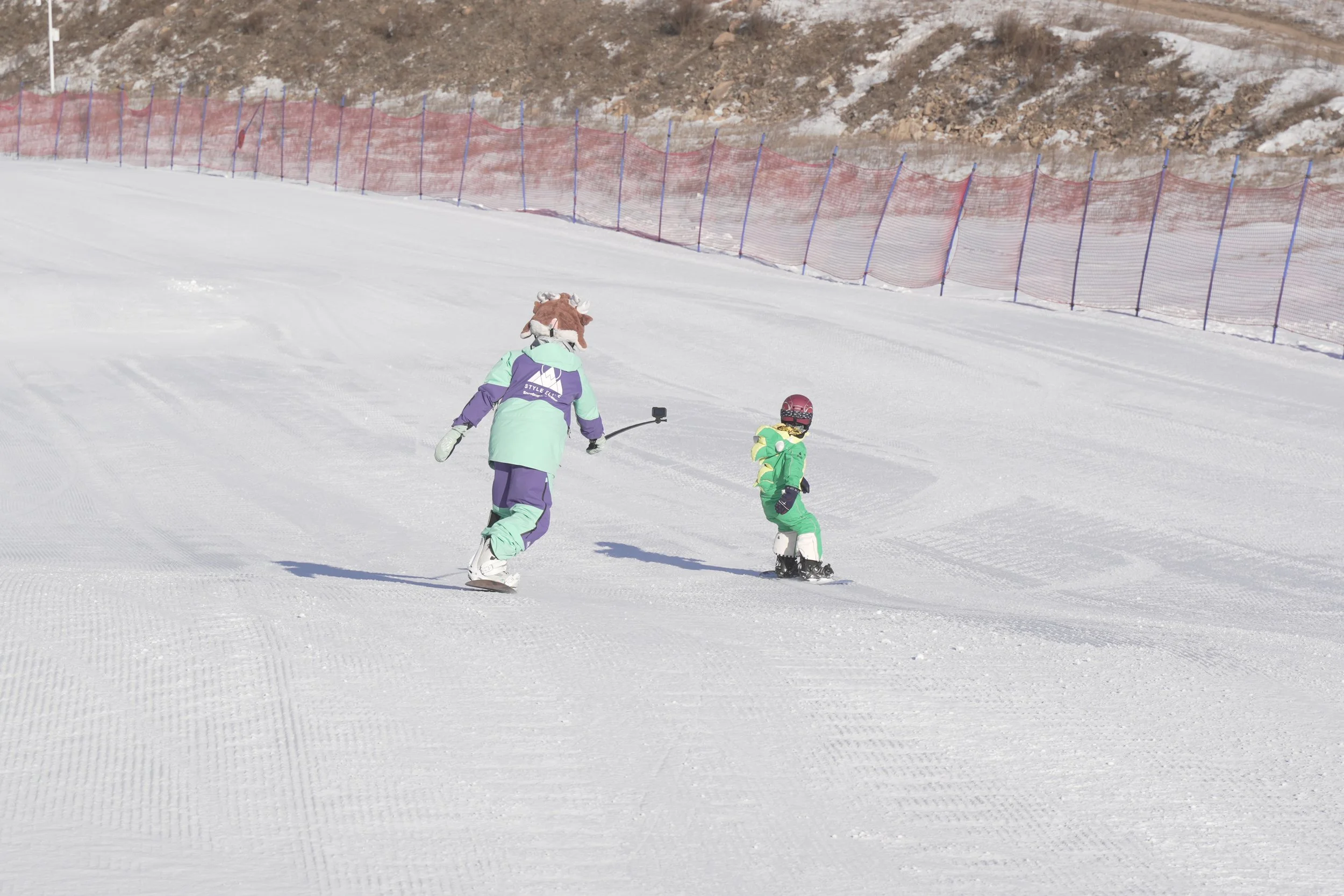 Two children in colorful ski outfits and helmets skiing on a snow-covered slope with a safety fence in the background.