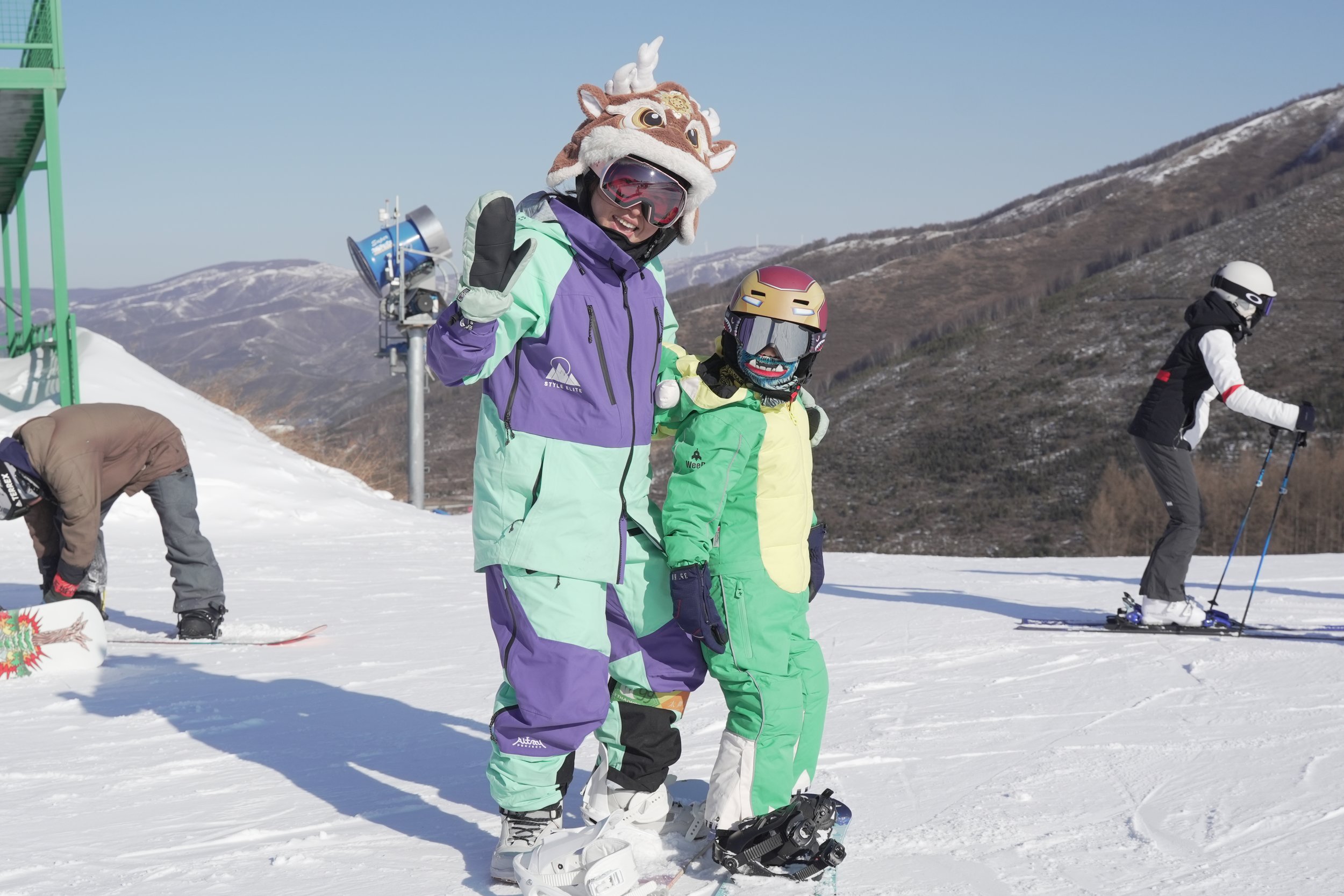 Two children in colorful ski outfits and protective helmets, one with a tiger hat, standing on snow with ski equipment, smiling and making a peace sign, with two other skiers in the background at a mountain ski resort.