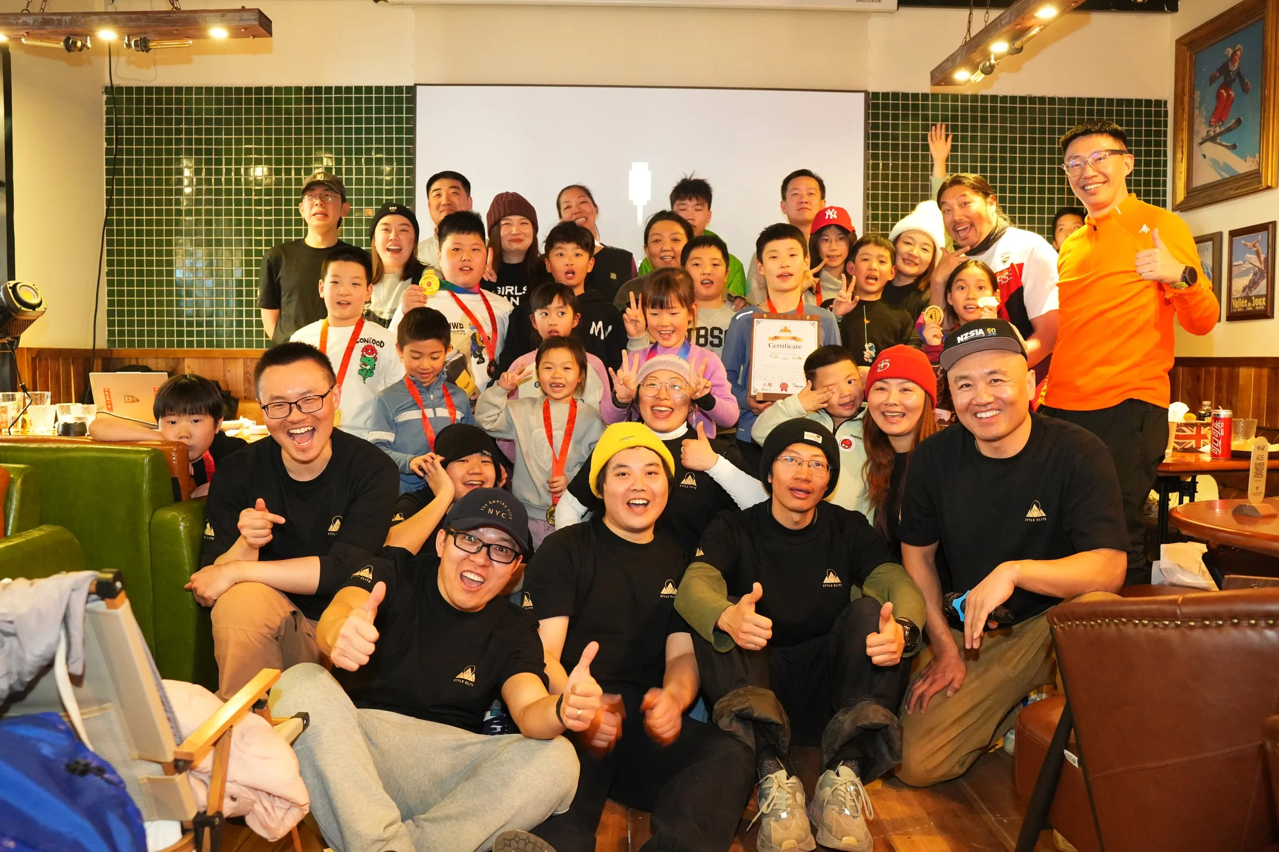 Group photo of children and adults celebrating indoors, some holding certificates and medals, with people smiling and making gestures like thumbs up and peace signs.
