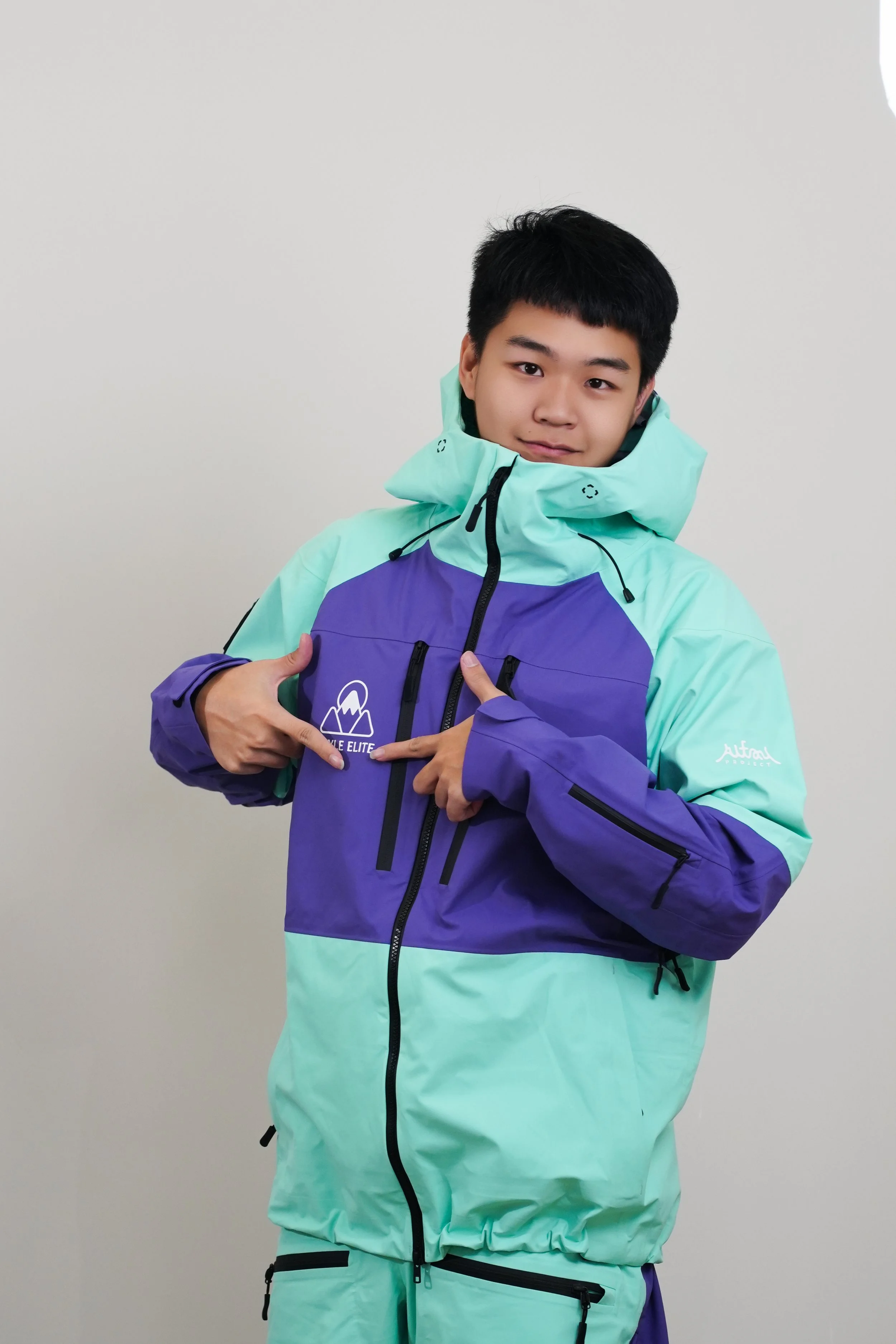A young person dressed in colorful skiing gear, including a helmet and goggles, standing on a snowy ski slope with ski lifts and mountains in the background.