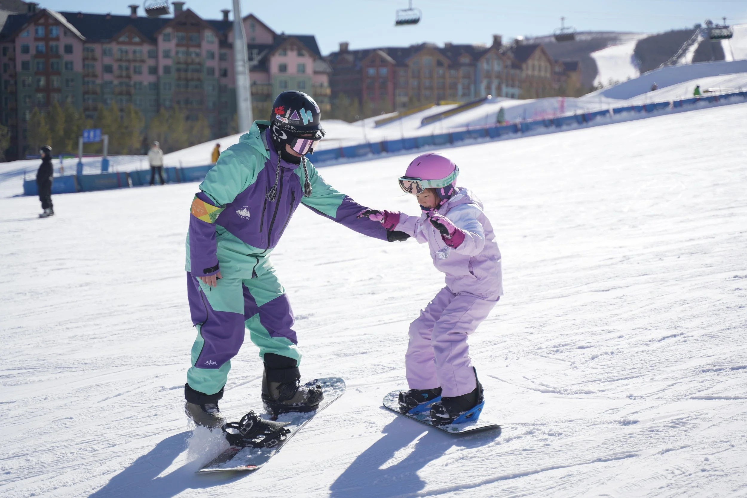 Two people on snowboards, an adult and a child, are snowboarding on a snowy slope. The adult is helping or instructing the child, holding the child's hand. The adult is dressed in a colorful ski jacket and pants, wearing a helmet and goggles. The child is dressed in a light pink snowsuit with a matching helmet and gloves, standing on a snowboard. In the background, other skiers and snowboarders are visible, with buildings and ski lifts in the distance.