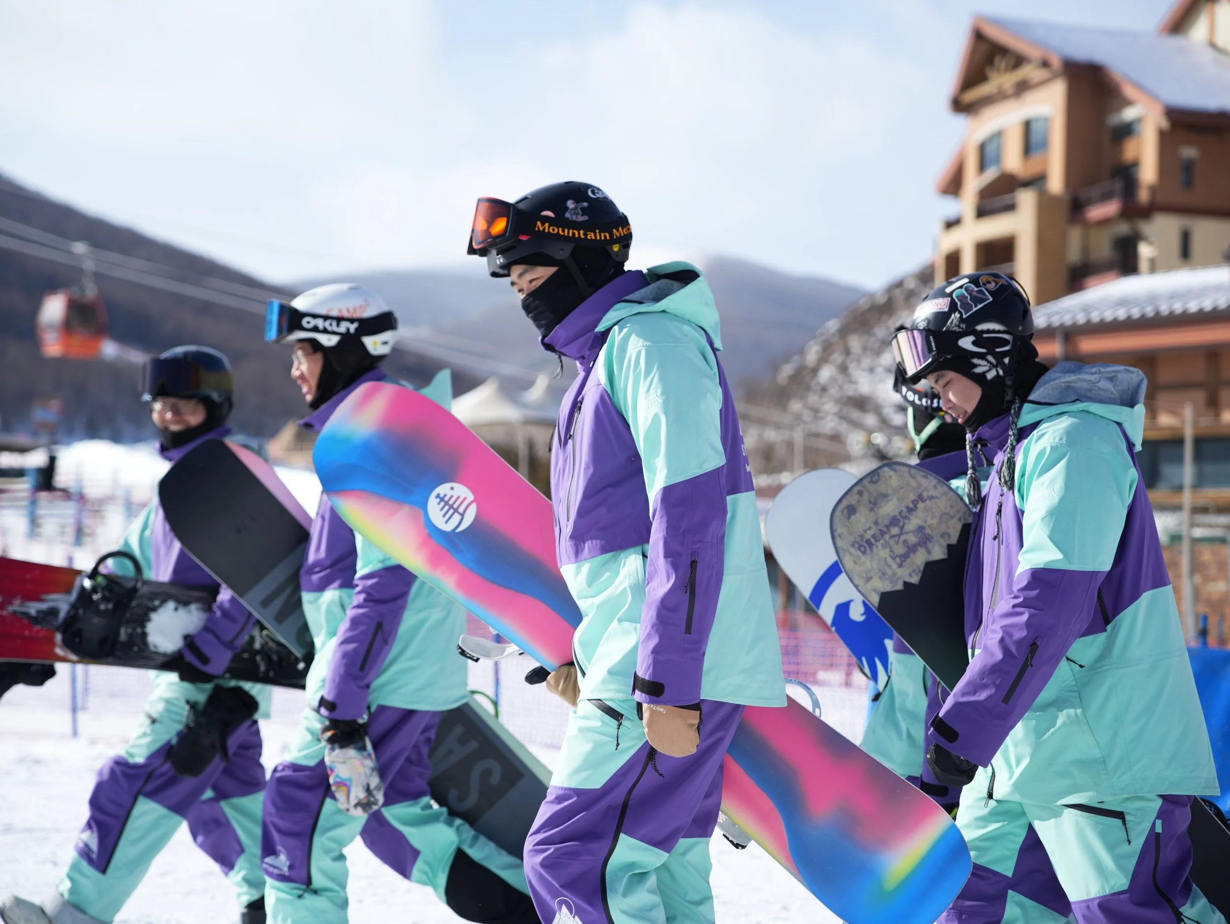 Group of four snowboarders in colorful winter gear with snowboards, walking on snow-covered ground at ski resort with chairlifts and cabins in background.