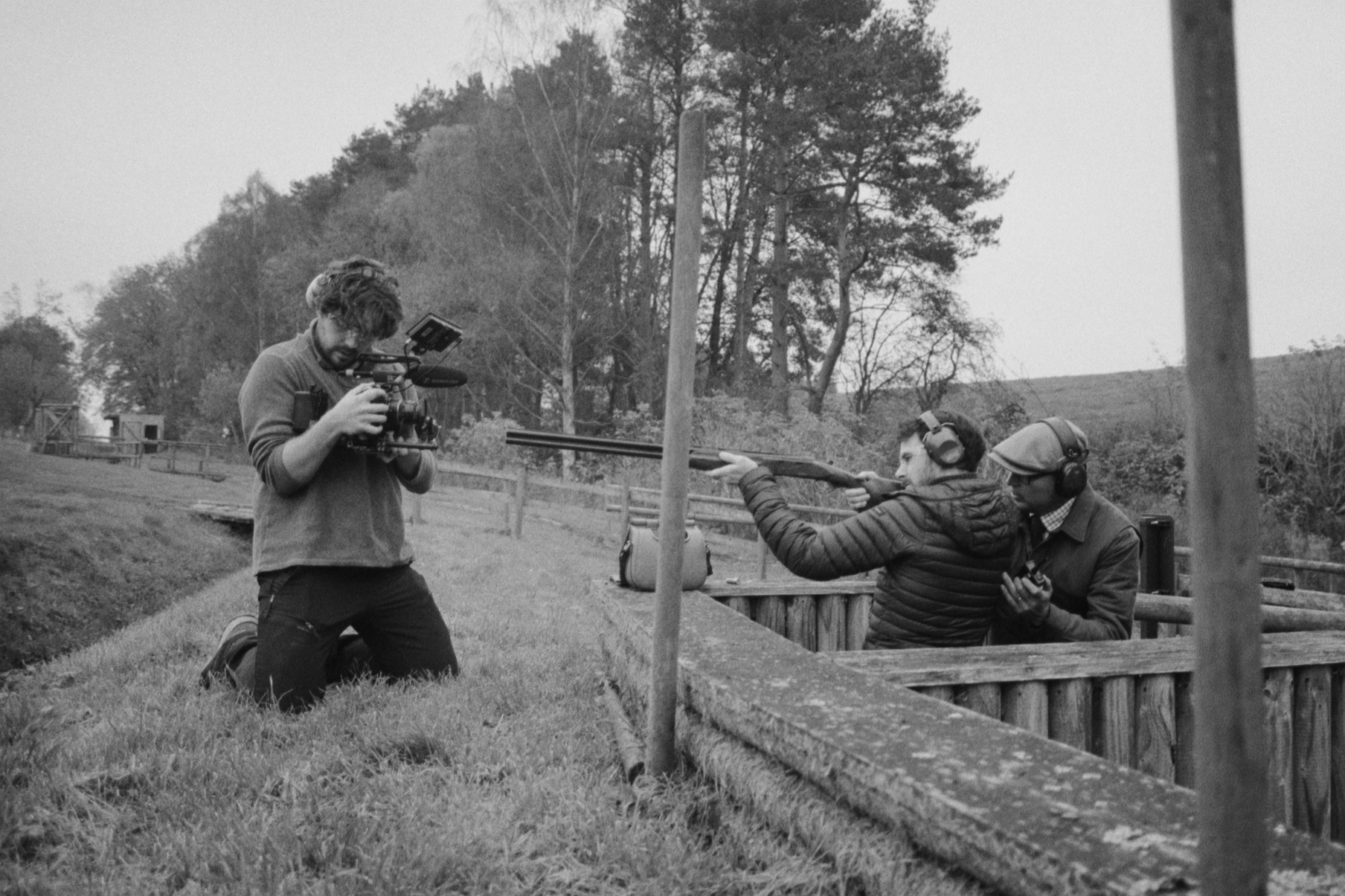 Two men, wearing headphones and jackets, are shooting a rifle at a rifle range while a cameraman films them. The scene is outdoors with trees and a fence in the background.