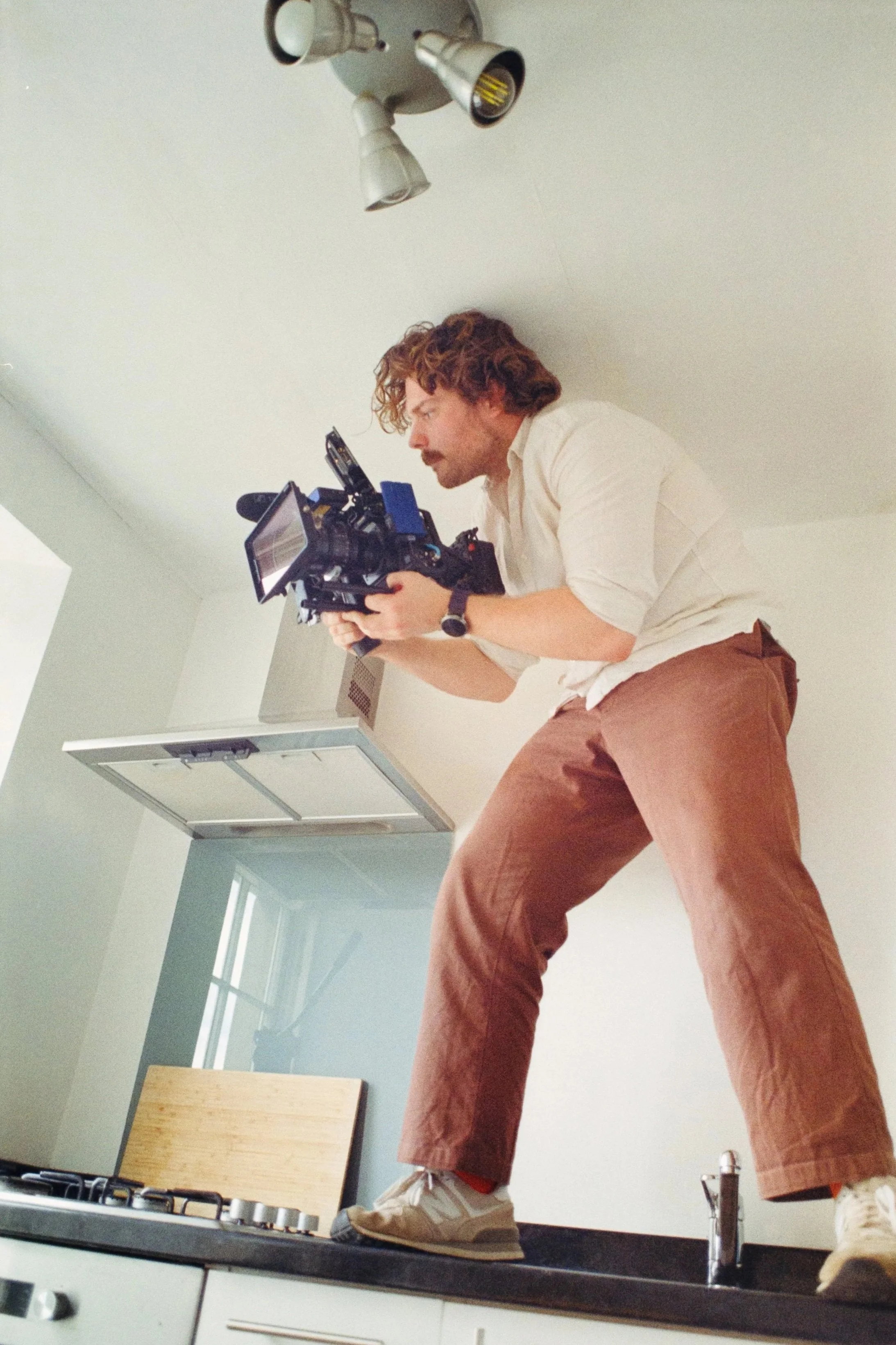 A man with curly hair, wearing a beige shirt and brown pants, standing on a kitchen counter, holding a professional camera and looking through its viewfinder.