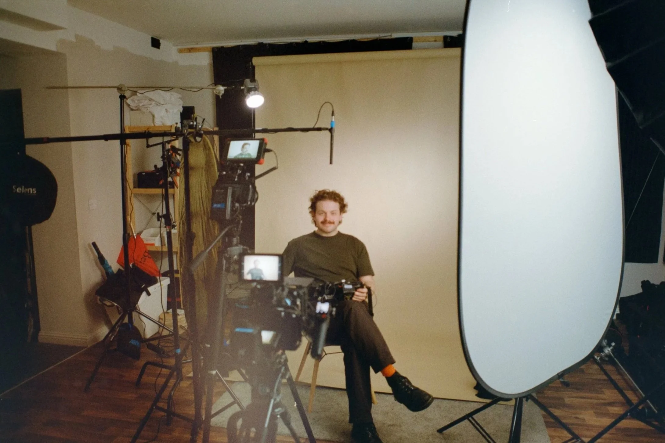 A man with curly hair and a mustache sitting on a stool in front of a beige backdrop during a photo or video shoot. Various photography equipment, including cameras, lights, and a large white reflector, are set up around him.