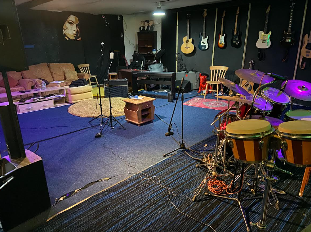 Music rehearsal or recording studio with guitars on the wall, musical instruments, microphones, and a drum set facing a couch and chairs.