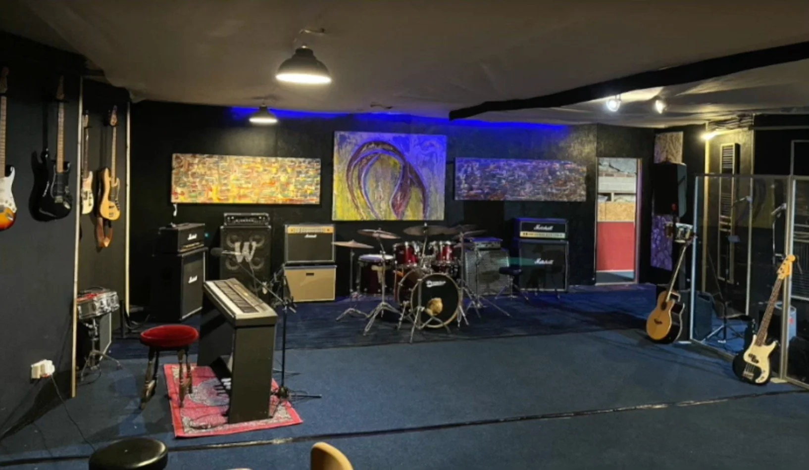 Empty music rehearsal studio with guitars hanging on the black wall, a drum set at the center, amplifiers, keyboards, and two paintings on the background wall, with some stage lights and a small window.