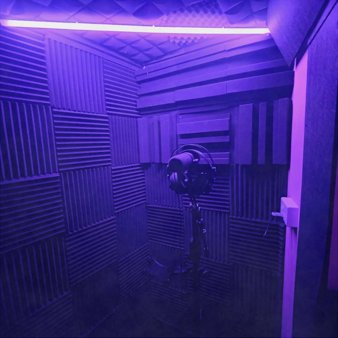 A soundproof recording booth with purple lighting, acoustic foam panels on the walls, a pair of headphones hanging from a stand, and a small white outlet on the wall.