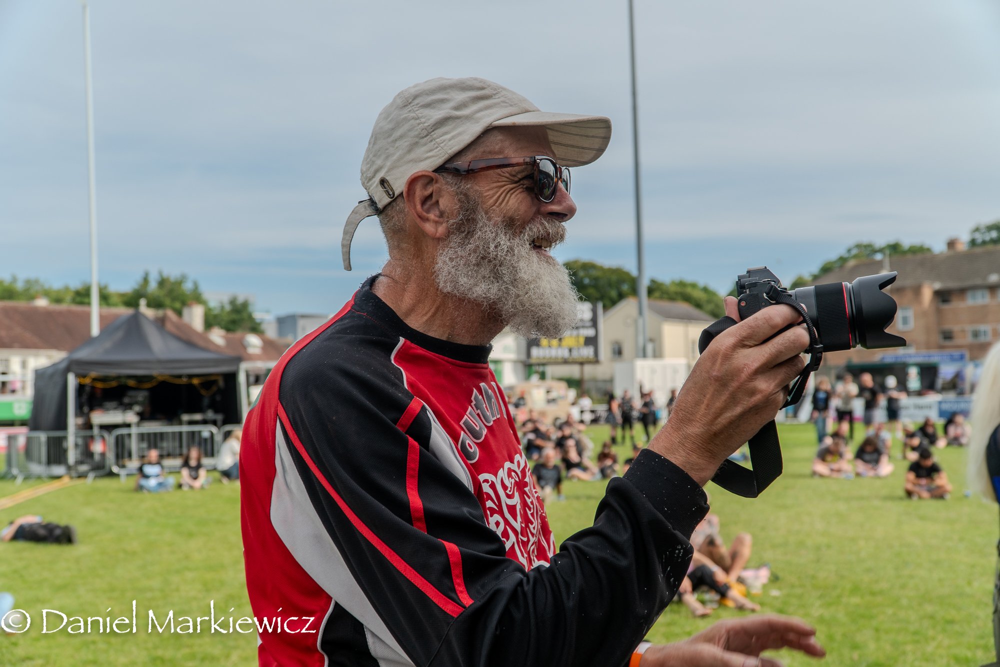 An elderly man with a gray beard wearing a beige cap, sunglasses, and a red and black sports jersey, holding a camera at an outdoor event with people sitting and standing on a grassy field, some tents, and buildings in the background.