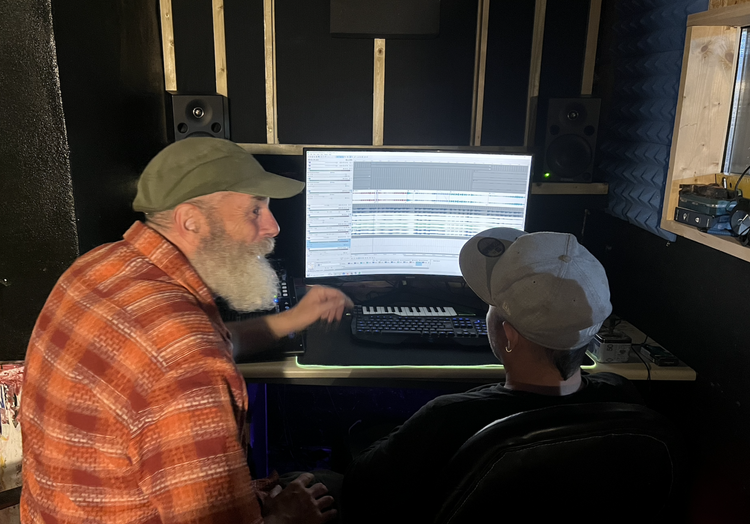 Two men with hats and glasses working on music production in a recording studio, looking at a large computer screen displaying digital audio tracks.