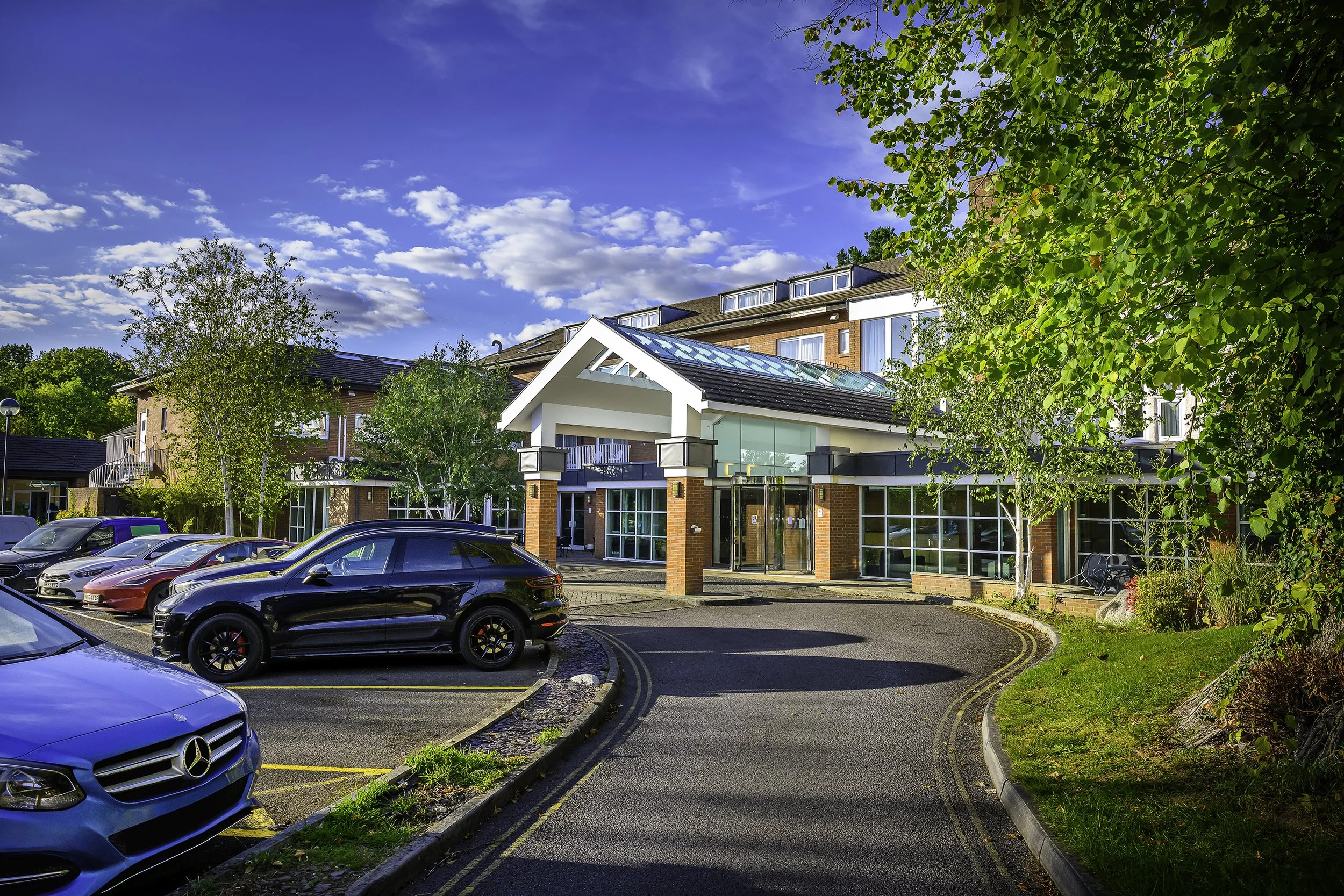 Exterior view of a modern residential building with a parking lot in front, surrounded by trees and greenery on a sunny day.