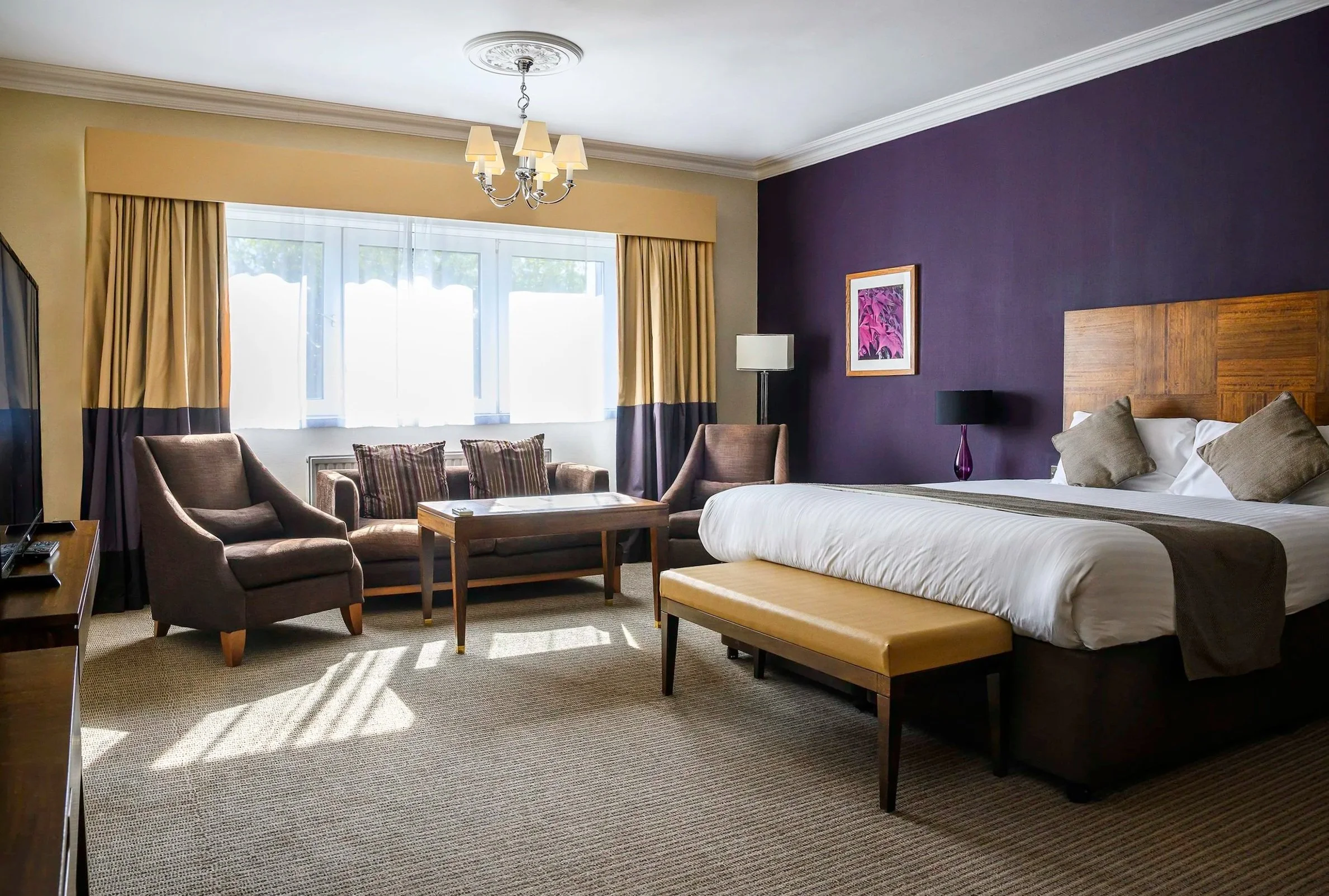 A hotel room with a large bed, a seating area with two armchairs and a sofa, a coffee table, and a window with curtains. The room has a purple accent wall, a framed picture, and a beige carpet.