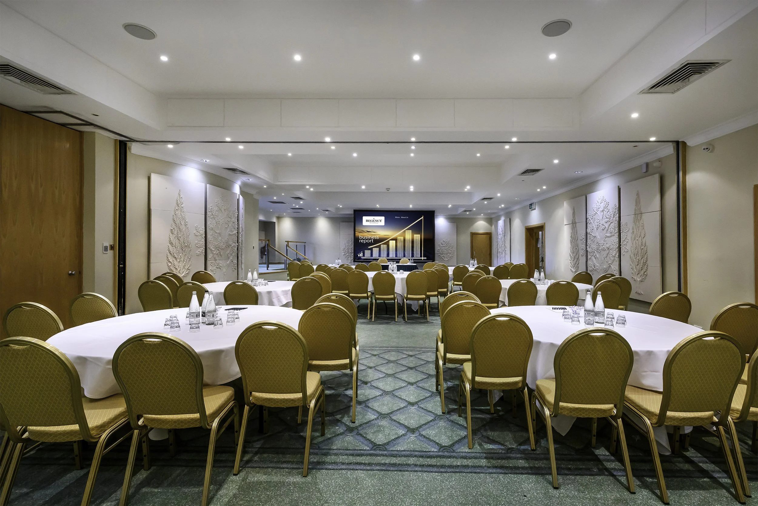 A conference room with round tables covered with white tablecloths, each set with water bottles and glasses. There are beige chairs around the tables. A large screen at the front displays a graph and the words "business report". The room has artwork on the walls and is well-lit with ceiling lights.