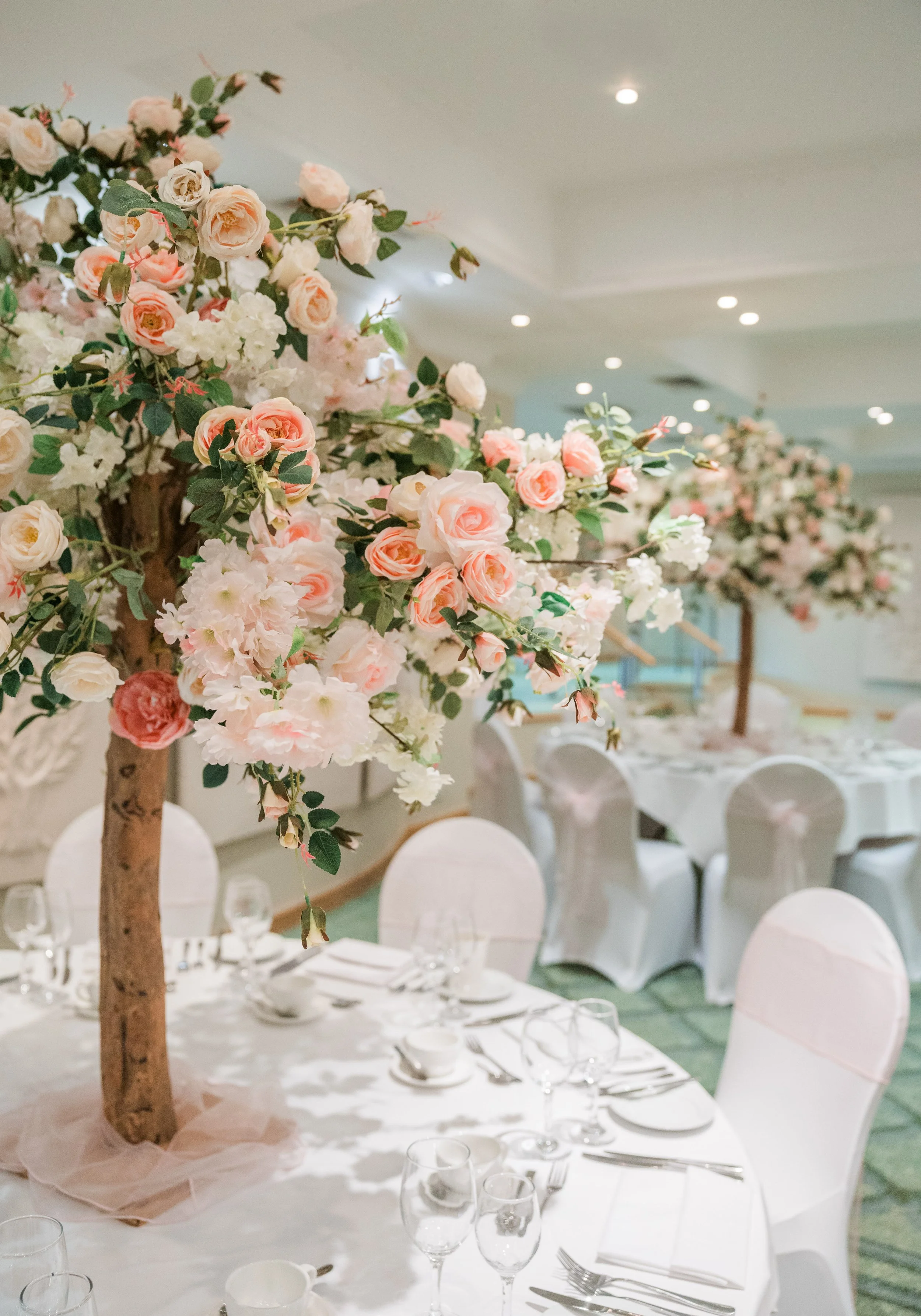 Elegant banquet hall with round tables decorated with white tablecloths, pink floral centerpieces on tall branches, and white chairs with covers, set for a formal event.