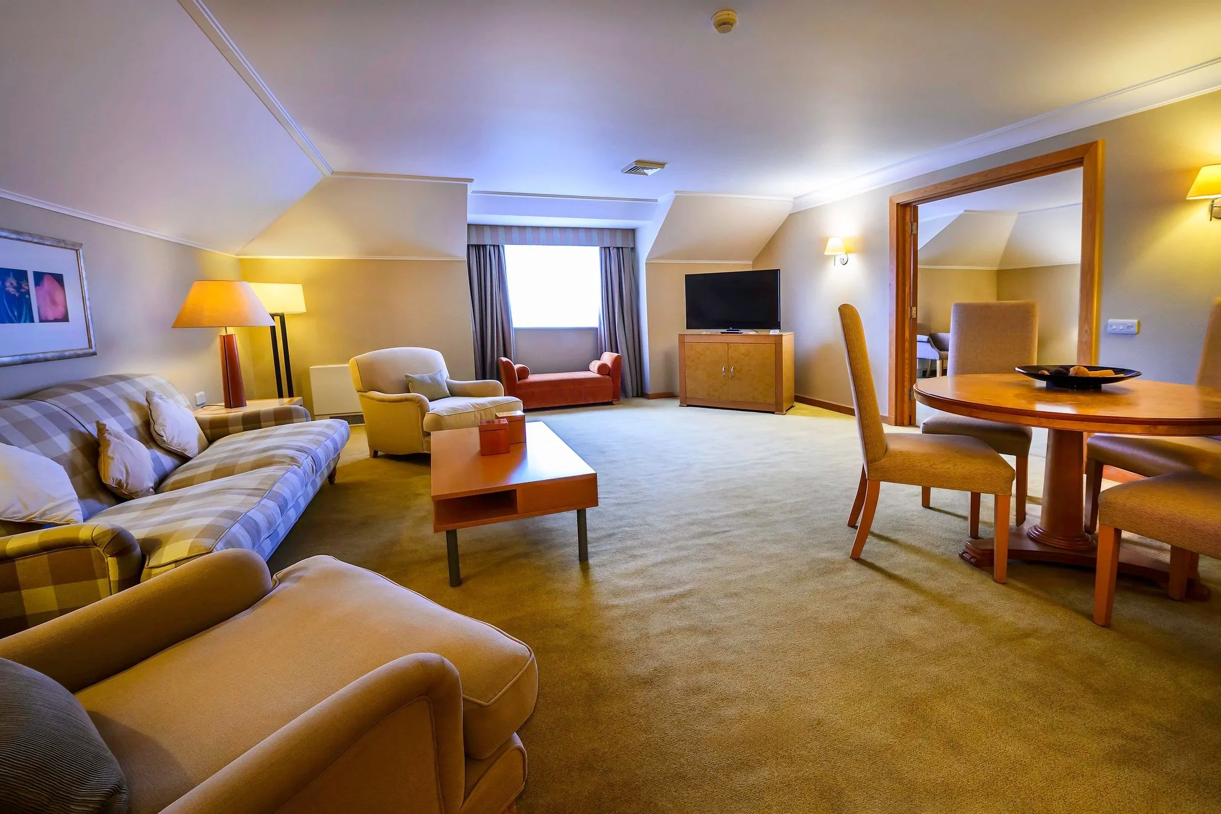 Spacious hotel living room with beige sofas, armchairs, a coffee table, dining table with chairs, TV, and window with curtains.