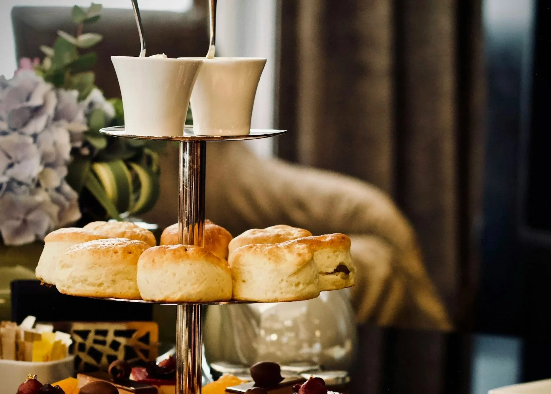 A tiered dessert stand with scones on the bottom and small cups on the top, set in a cozy indoor environment with flowers and curtains in the background.