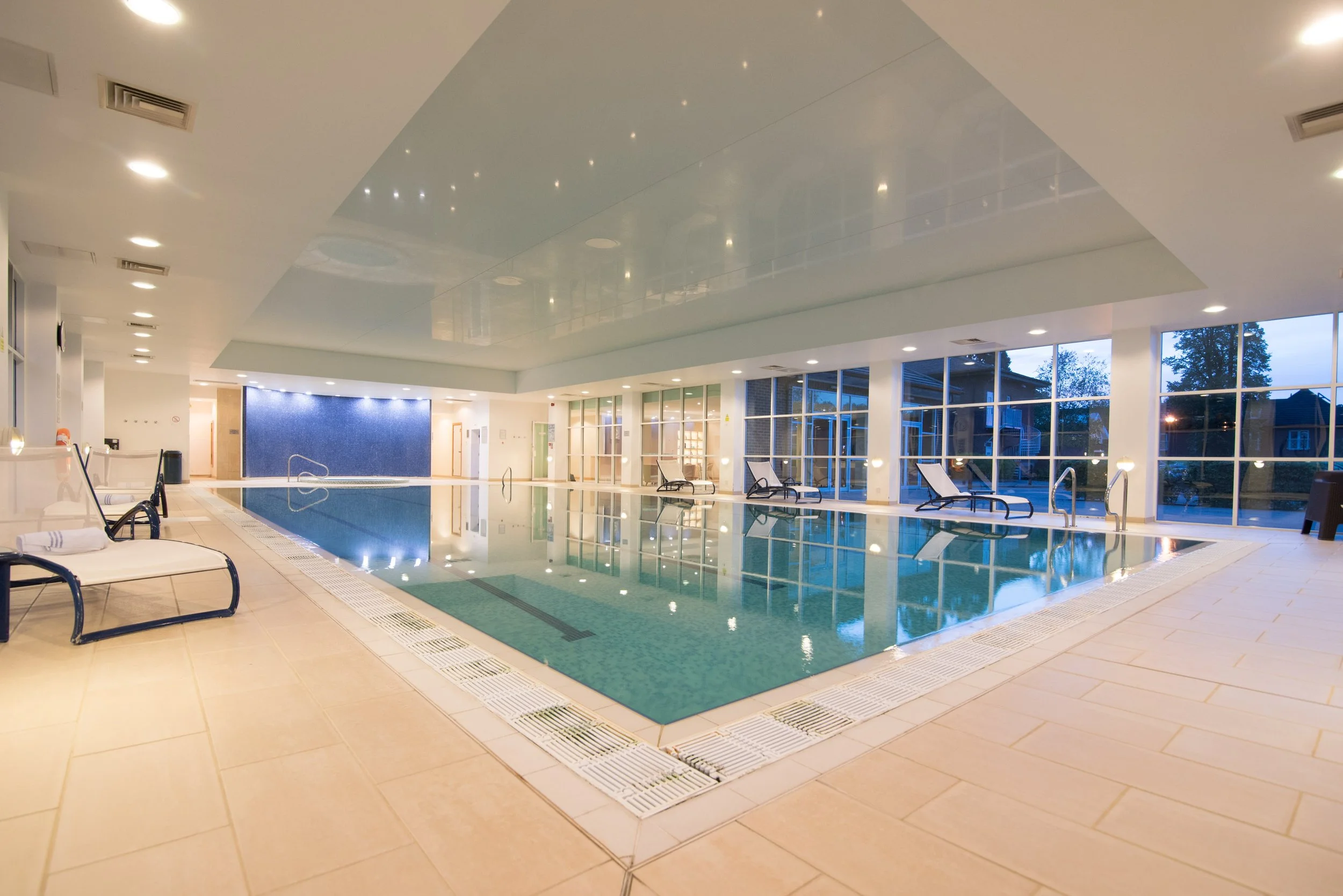 Indoor swimming pool with lounge chairs along the sides, large windows showing outside trees, and ambient ceiling lighting.