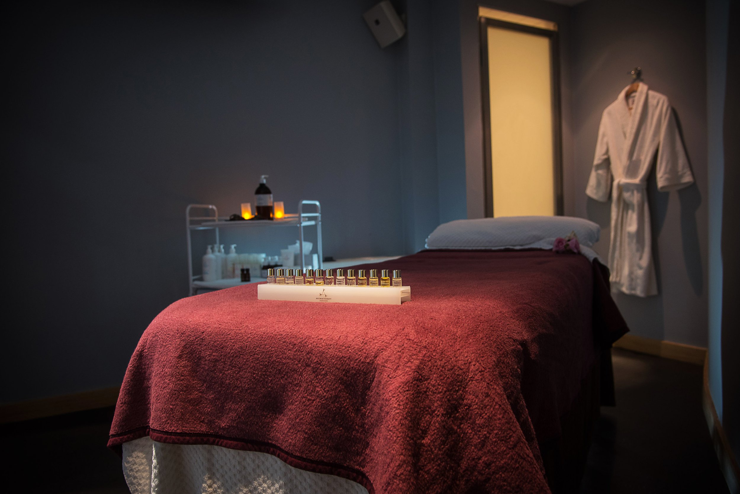 A massage room with a massage table covered in a burgundy blanket, scented oils arranged on a tray on the table, and a white robe hanging on the wall. Small candles are lit behind the table, creating a calming atmosphere.