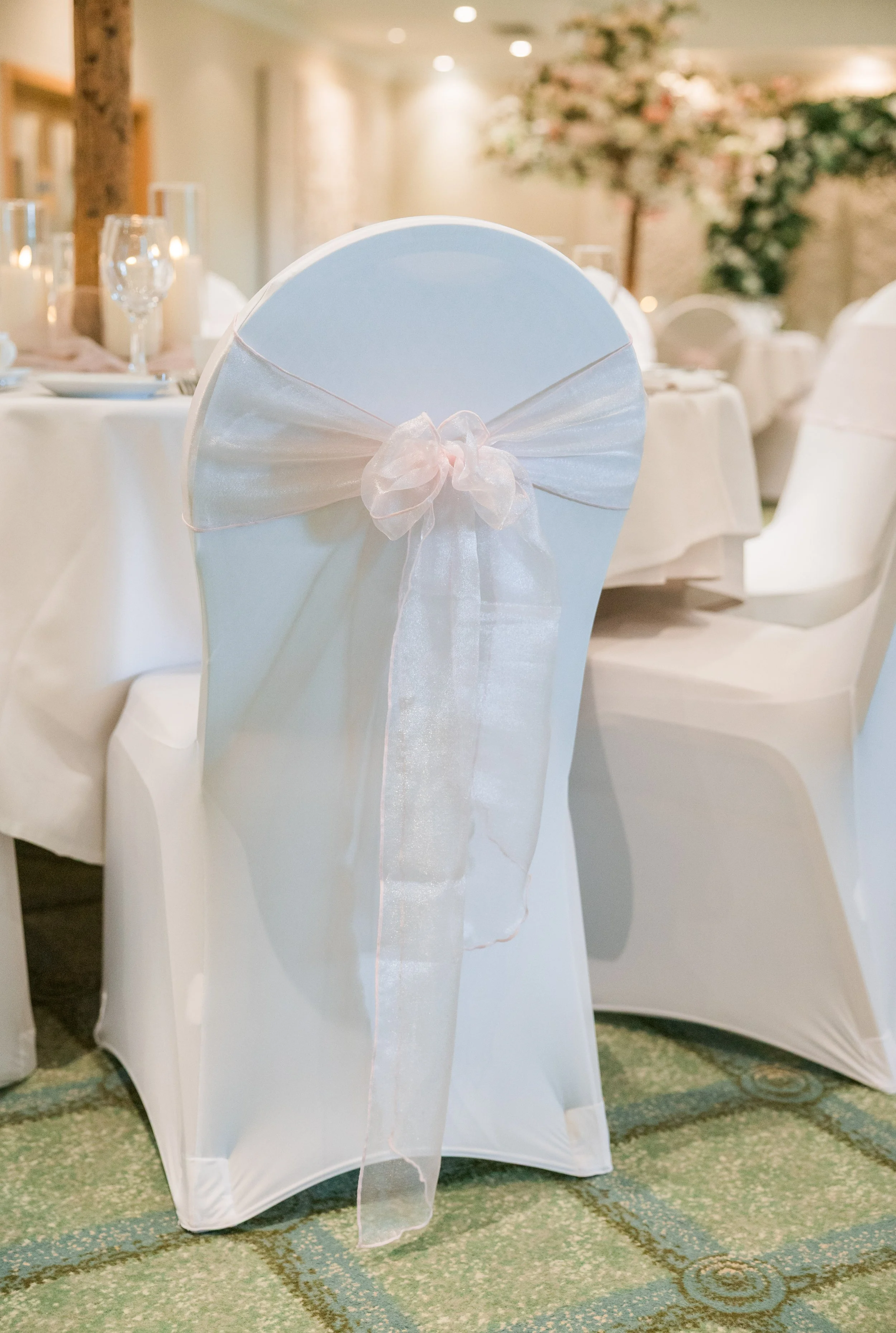 Wedding reception table with white chairs decorated with sheer ribbons tied in bows, set with glassware and candles, in a decorated hall with floral arrangements.