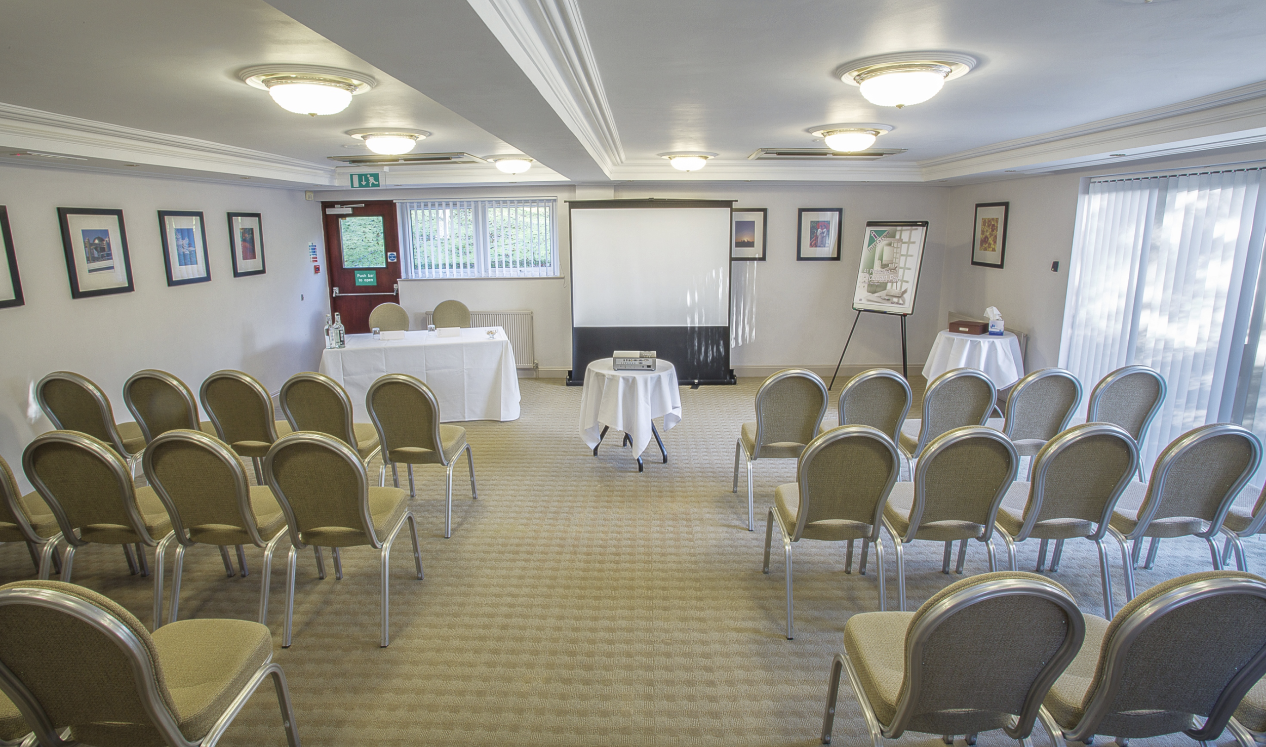 Meeting room set up for a presentation with chairs arranged in rows, a projector and screen at the front, and a table with chairs and bottled water at the side.