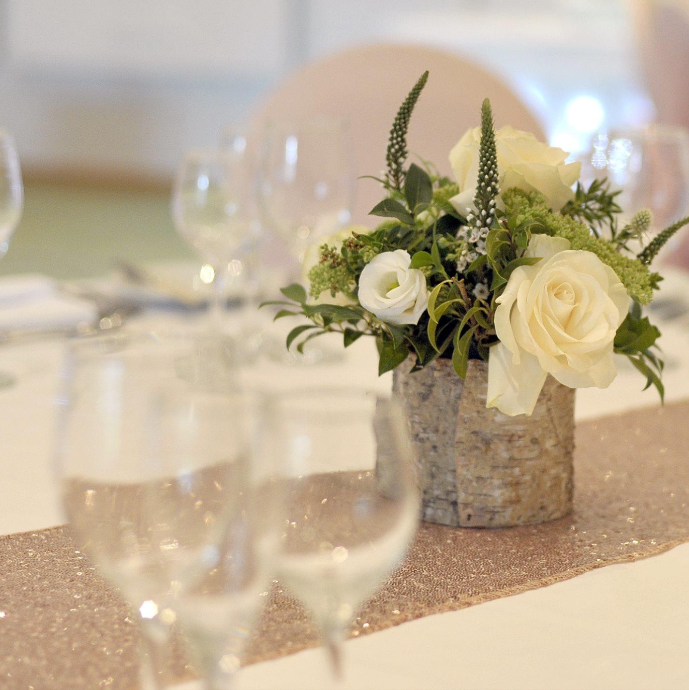 A floral centerpiece with white roses and greenery in a birch bark vase on a table with a glittering gold table runner.