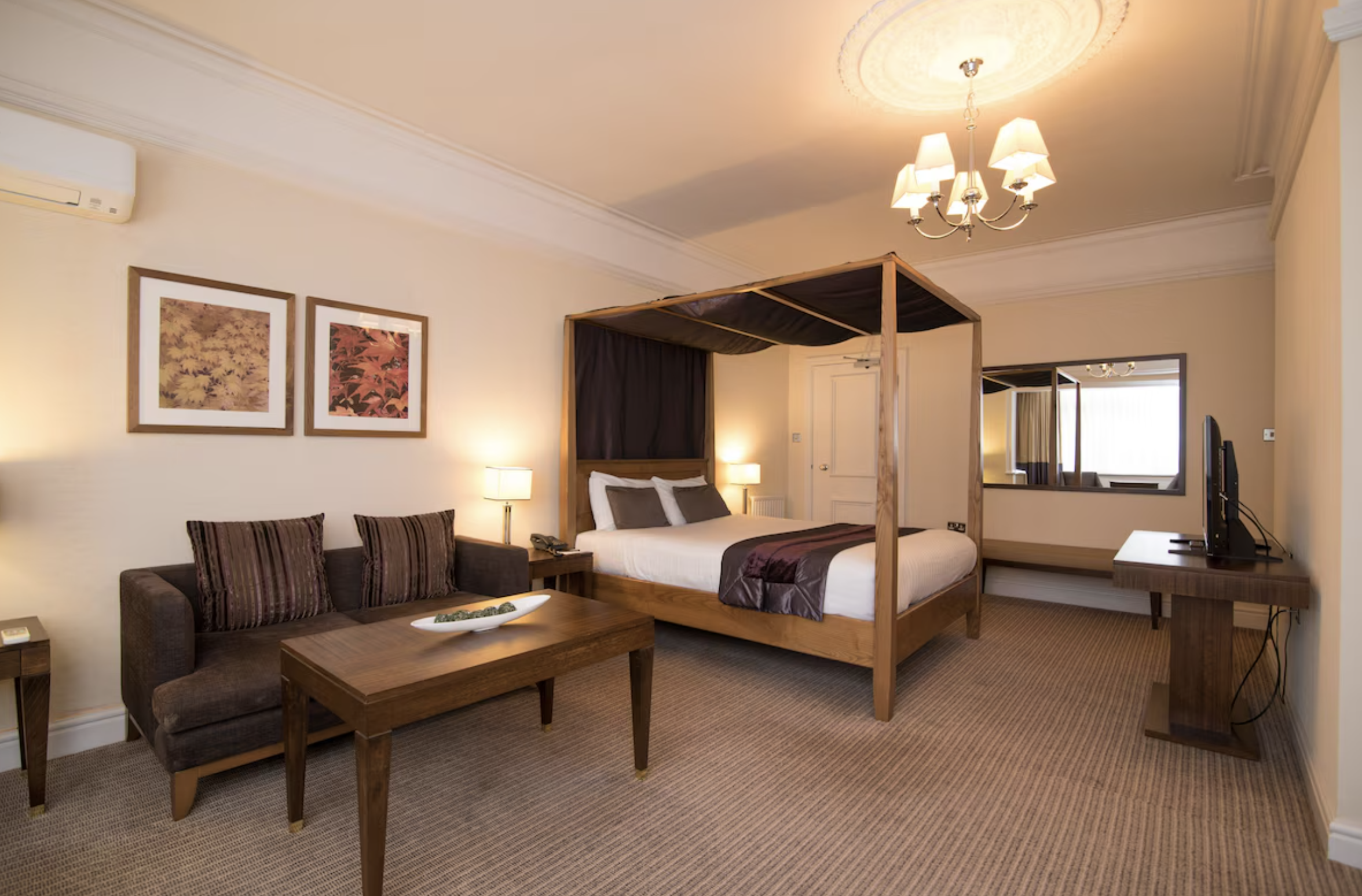 A hotel room with a four-poster bed, a brown sofa with striped pillows, a wooden coffee table, a television on a wooden stand, and two bedside lamps. The room has beige walls, a chandelier, and framed artwork on the wall.