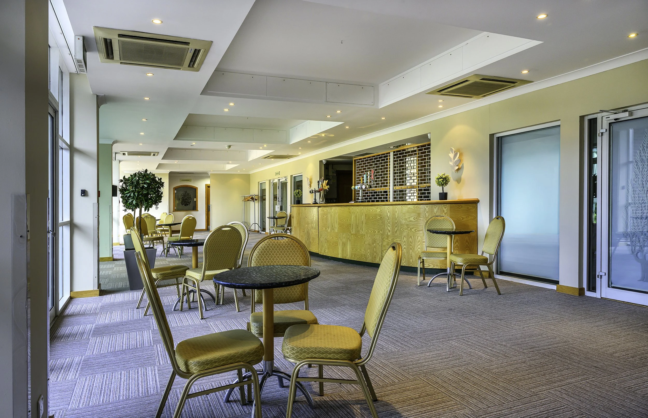 Empty indoor room with yellow chairs, round tables, and a bar counter, illuminated by ceiling lights and natural light from windows.