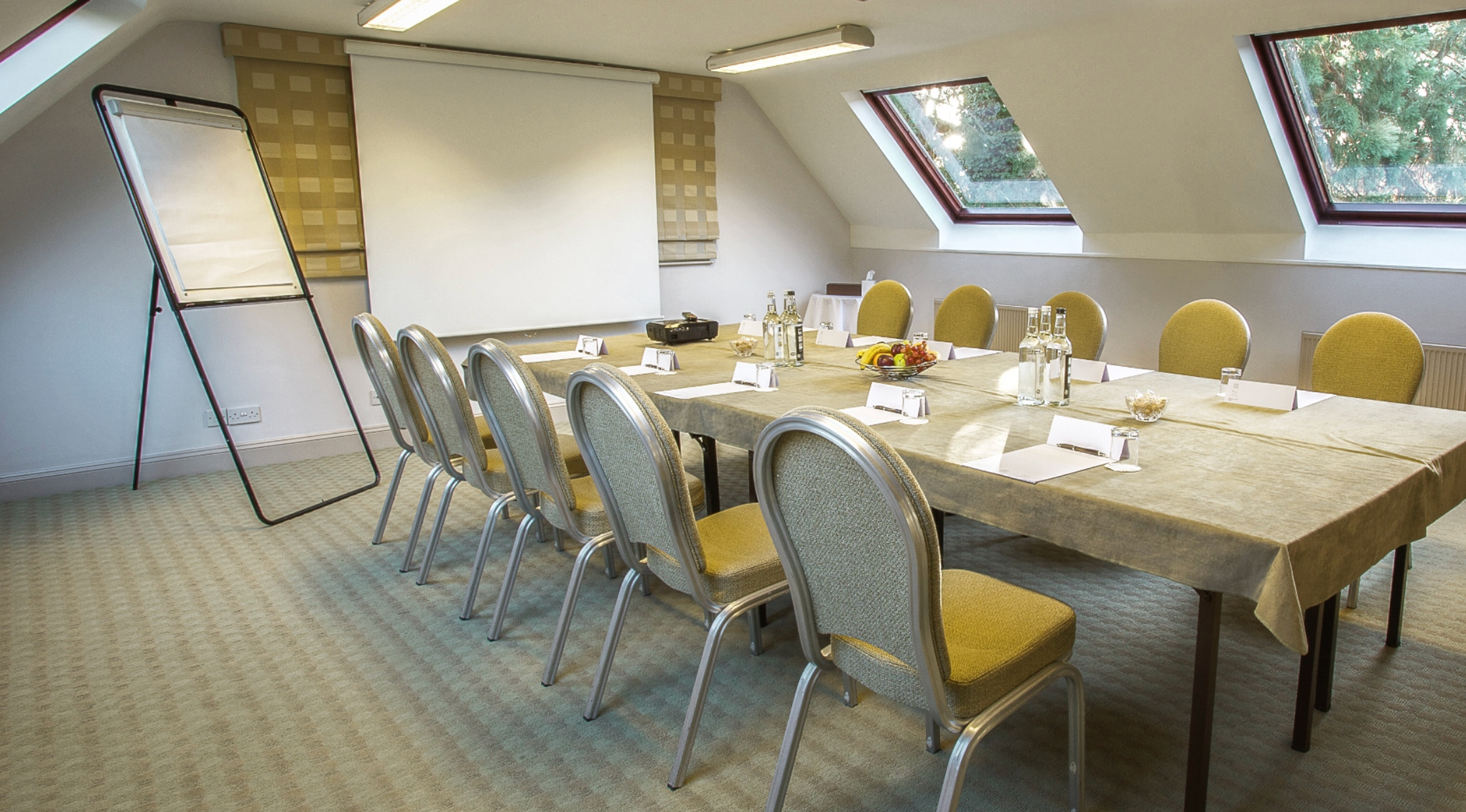 A conference room with a long table set with bottled water, glasses, and notepads, surrounded by yellow chairs, with large skylight windows and a whiteboard.
