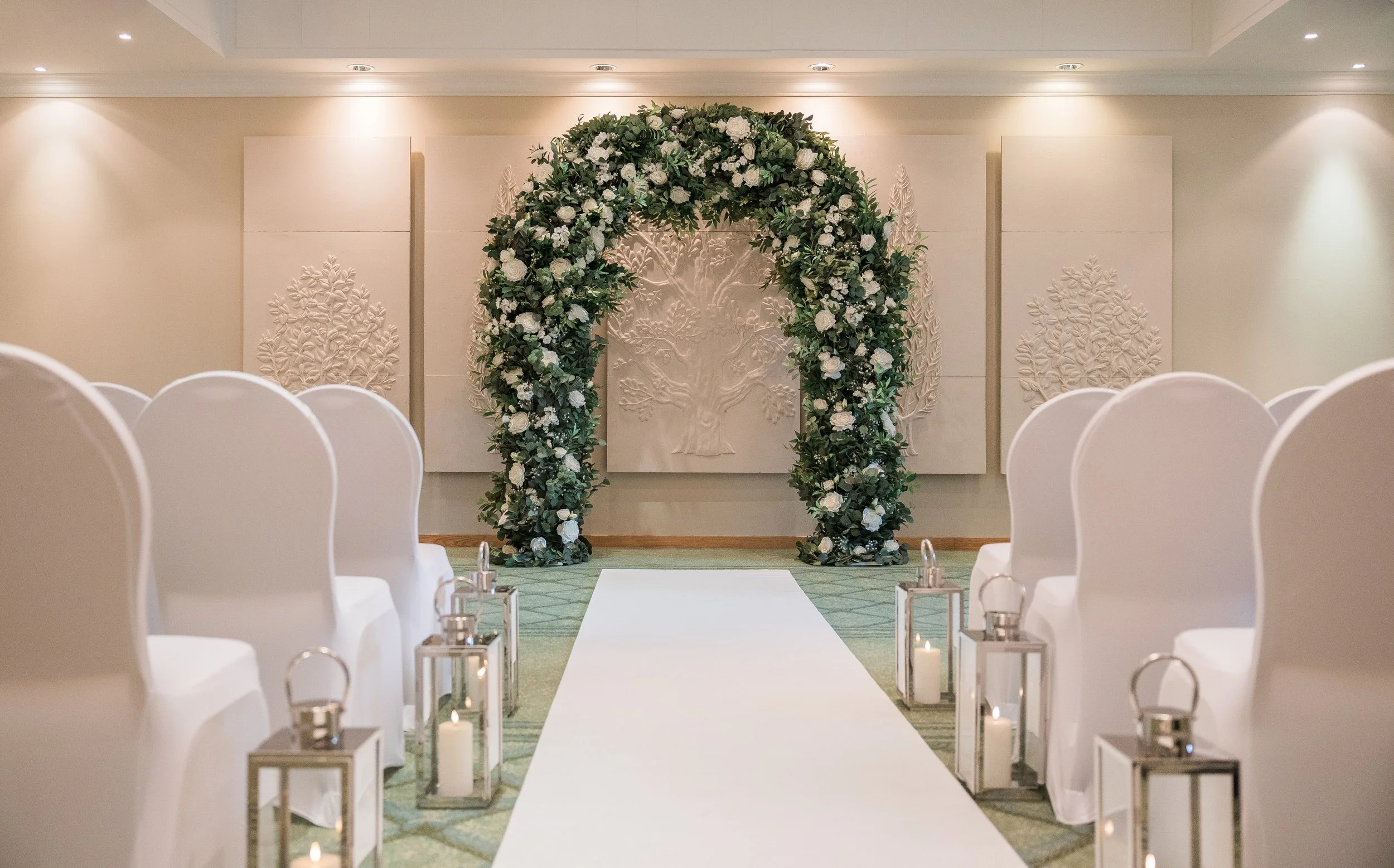 Indoor wedding ceremony setup with a white aisle runner, white chairs, floral arch, and lanterns with candles along the aisle.