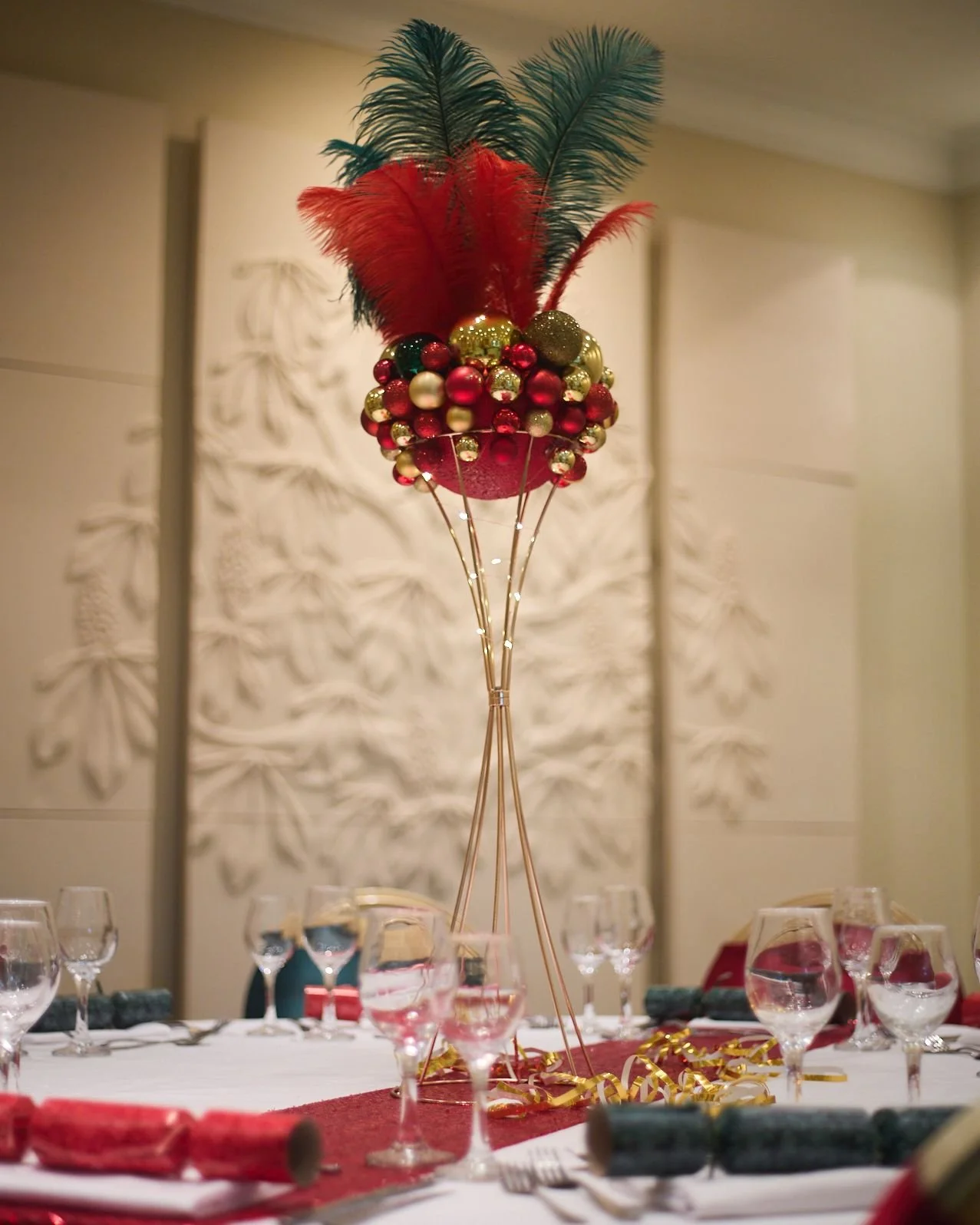 Festive table centerpiece with red and green feathers, gold and red ornaments, and string lights, surrounded by wine glasses and table settings for a holiday celebration.