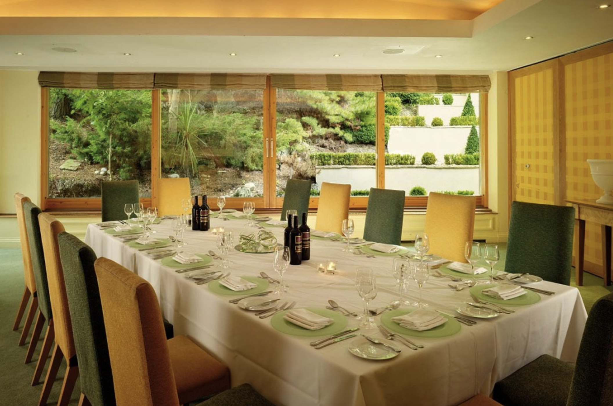 Large dining table set for a formal meal with wine glasses, plates, silverware, and candles, in a bright room with large windows overlooking a landscaped garden.