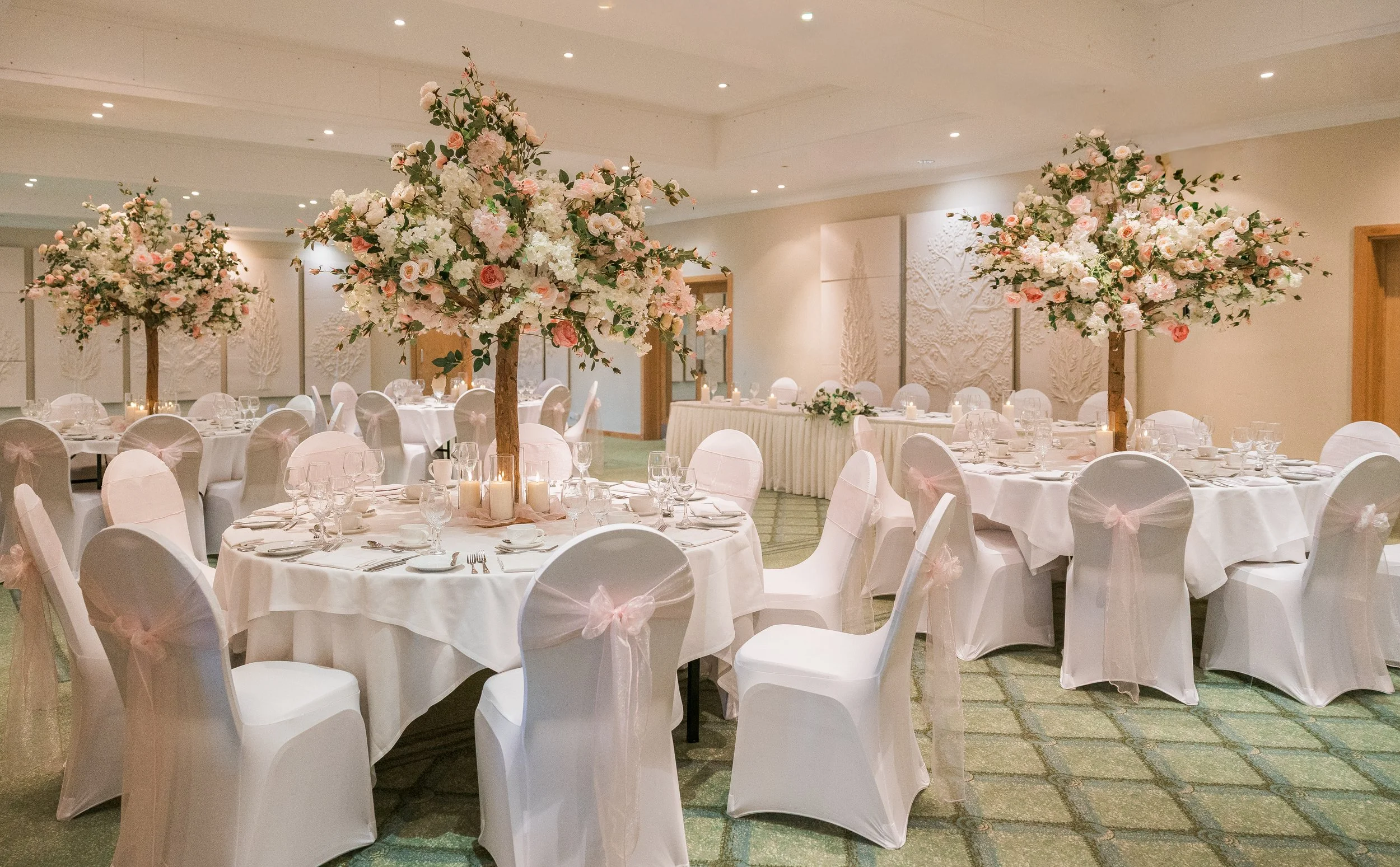 Elegant banquet hall decorated with large pink and white floral centerpieces on round tables covered with white tablecloths, white chairs with pink bows, and soft ambient lighting for a wedding or special event.