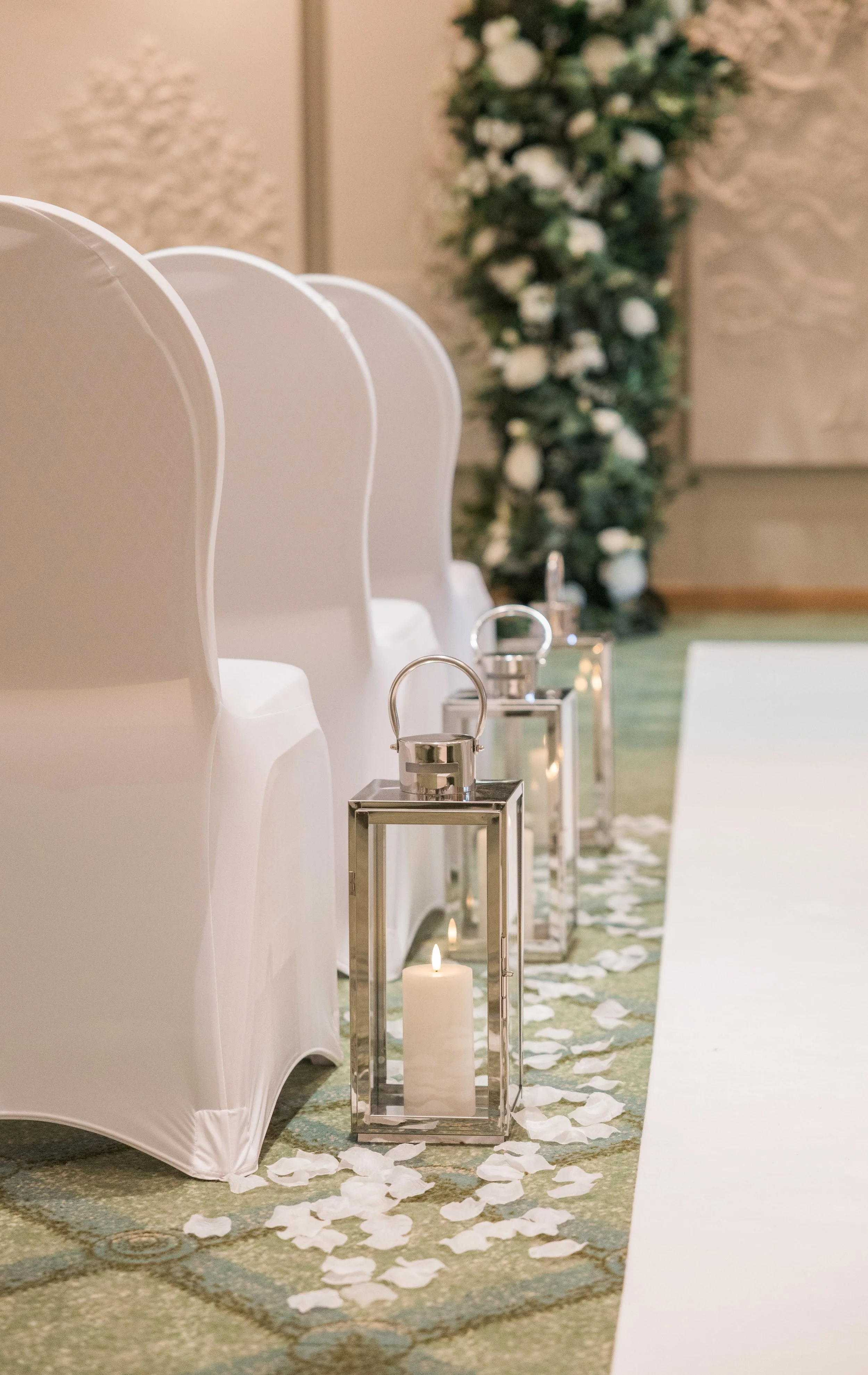 Wedding ceremony setup with white chairs, glass lanterns with lit candles, and flower petals on the aisle against a backdrop of floral arrangements.