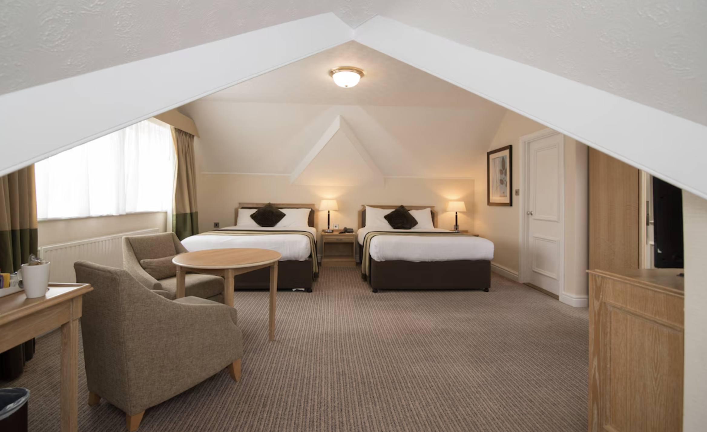 A spacious hotel room with two queen beds, nightstands with lamps, a sitting area with two armchairs and a round table, a window with curtains, and a wooden dresser with a TV, under a vaulted ceiling with a light fixture.