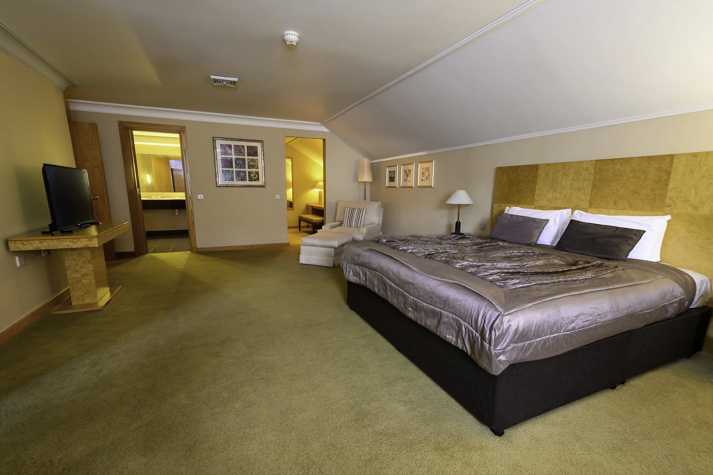 Hotel room with a large bed, beige walls, a chair with an ottoman, a lamp, and a flat-screen TV on a stand.