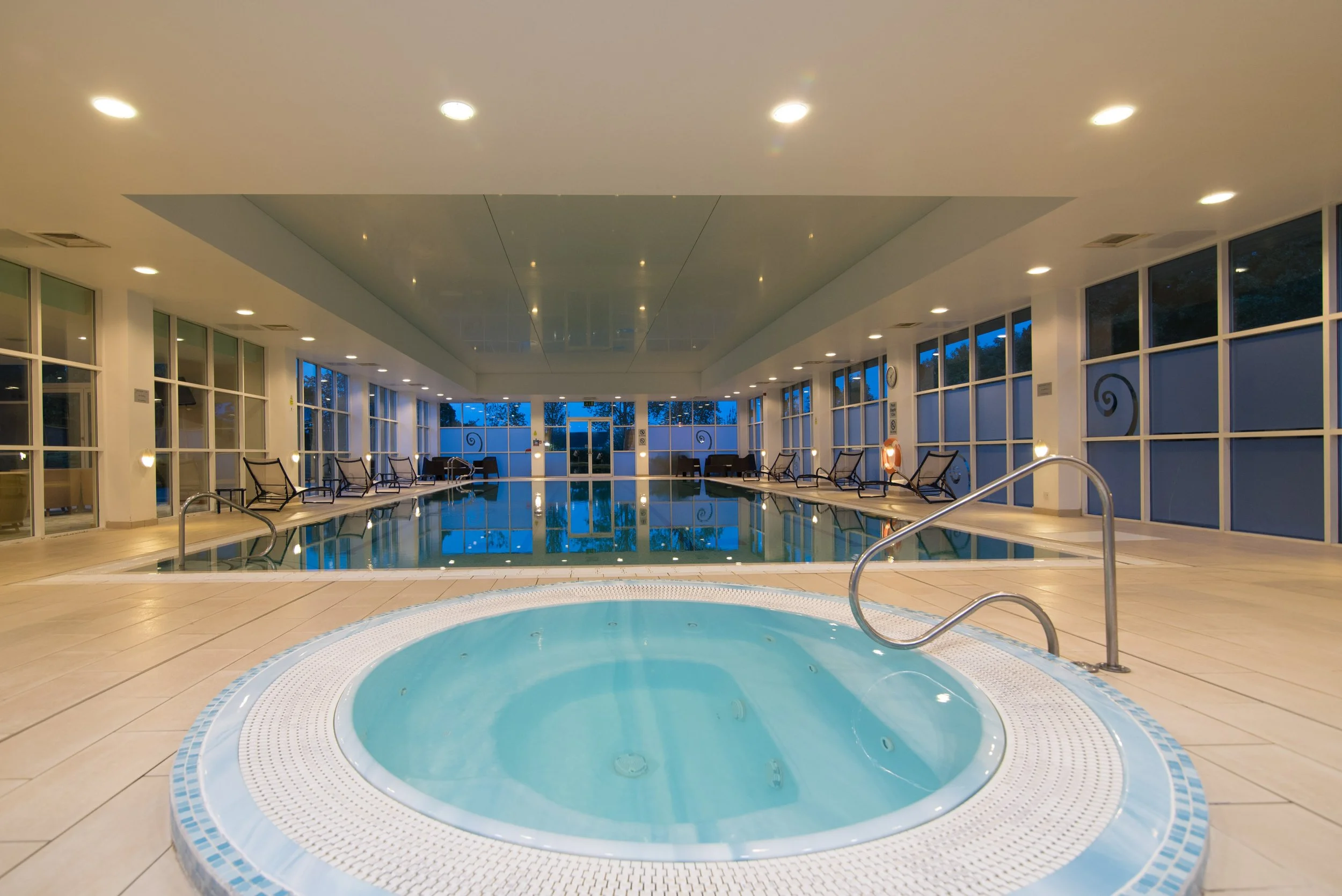 Indoor swimming pool with a hot tub in the foreground, lounge chairs along the sides, large windows, and ceiling lights.