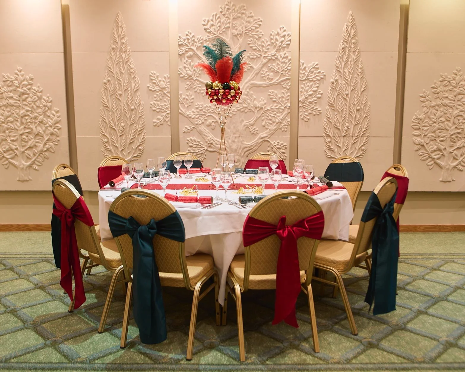 A banquet table set for a celebration with a colorful feather centerpiece. The table has white tablecloth, wine glasses, and napkins. Chairs around the table are decorated with red, blue, and black ribbons tied in bows.
