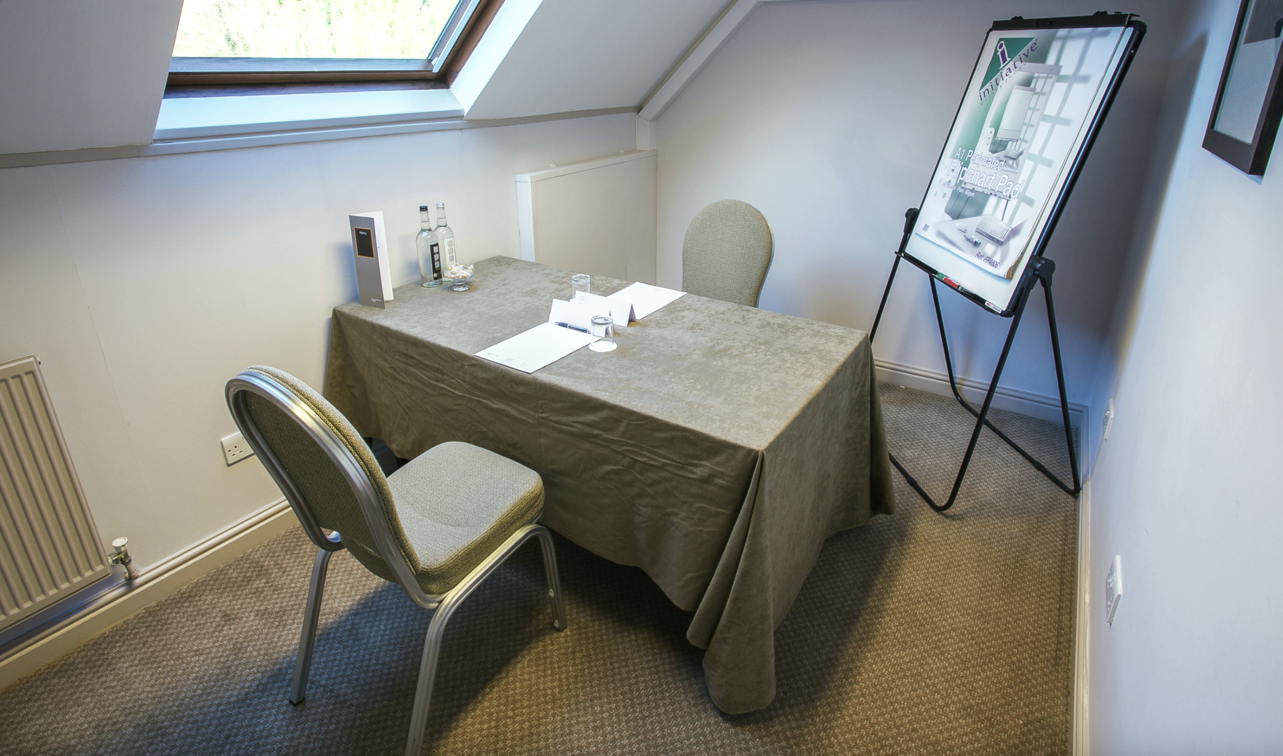 Small meeting room with a rectangular table covered in a brown tablecloth, two chairs, one with a green cushion, positioned around the table, a skylight providing natural light, a flip chart with a marketing presentation, water bottles, glasses, and a digital device on the table.