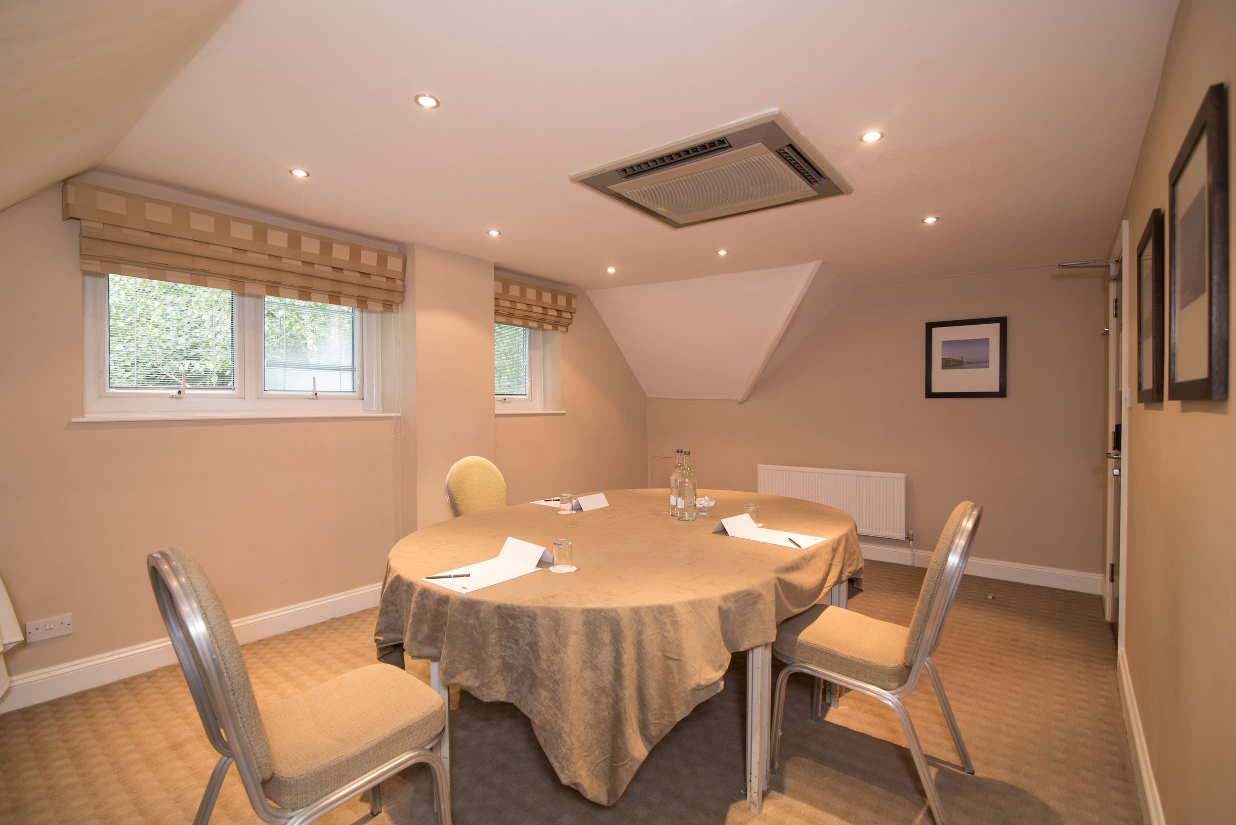 Large dining table set for a formal meal with wine glasses, plates, silverware, and candles, in a bright room with large windows overlooking a landscaped garden.