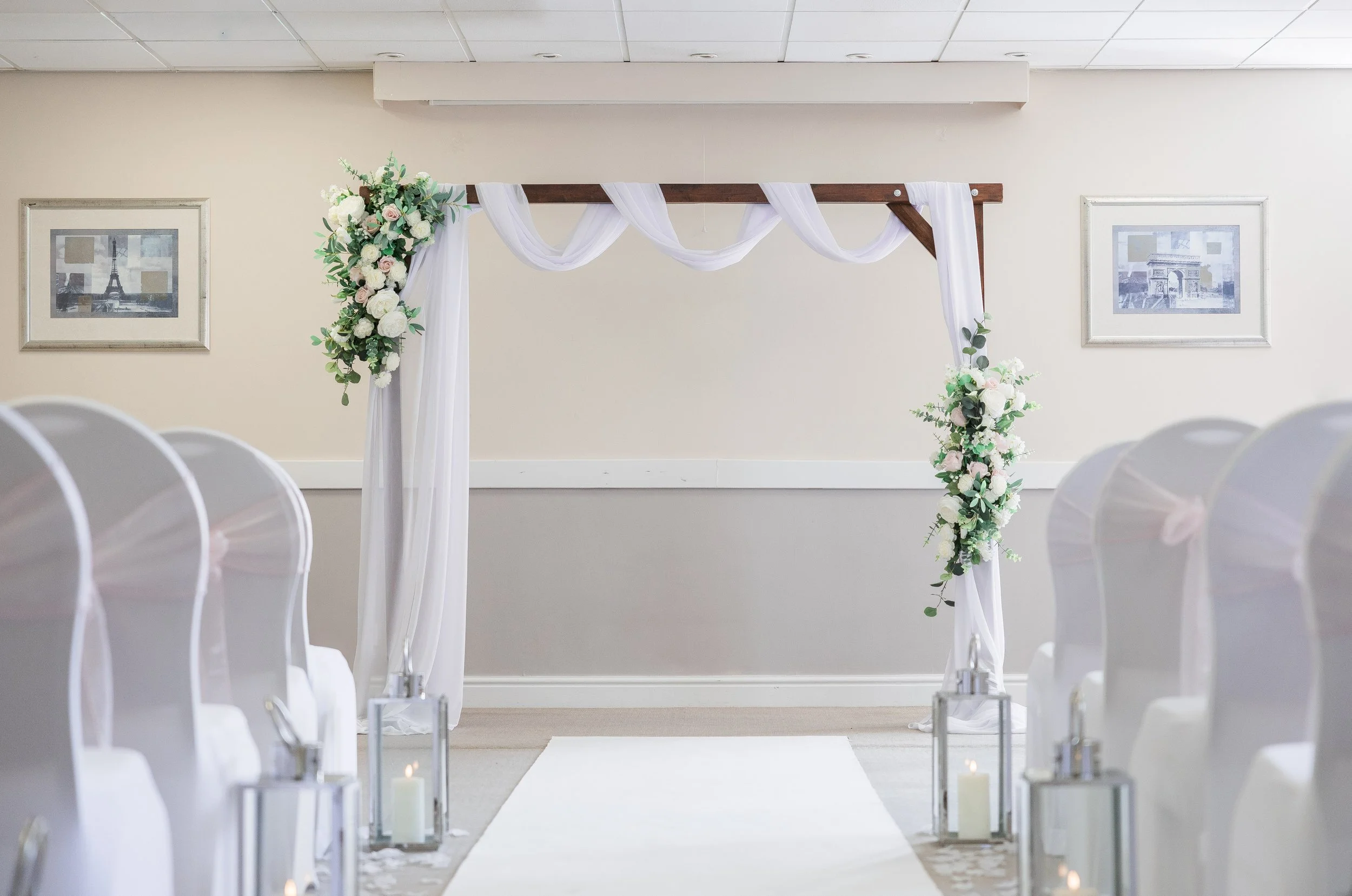 Wedding ceremony setup with a white aisle runner, lanterns with candles, white chairs with pink bows, and a wooden arch decorated with white and pink flowers and greenery.