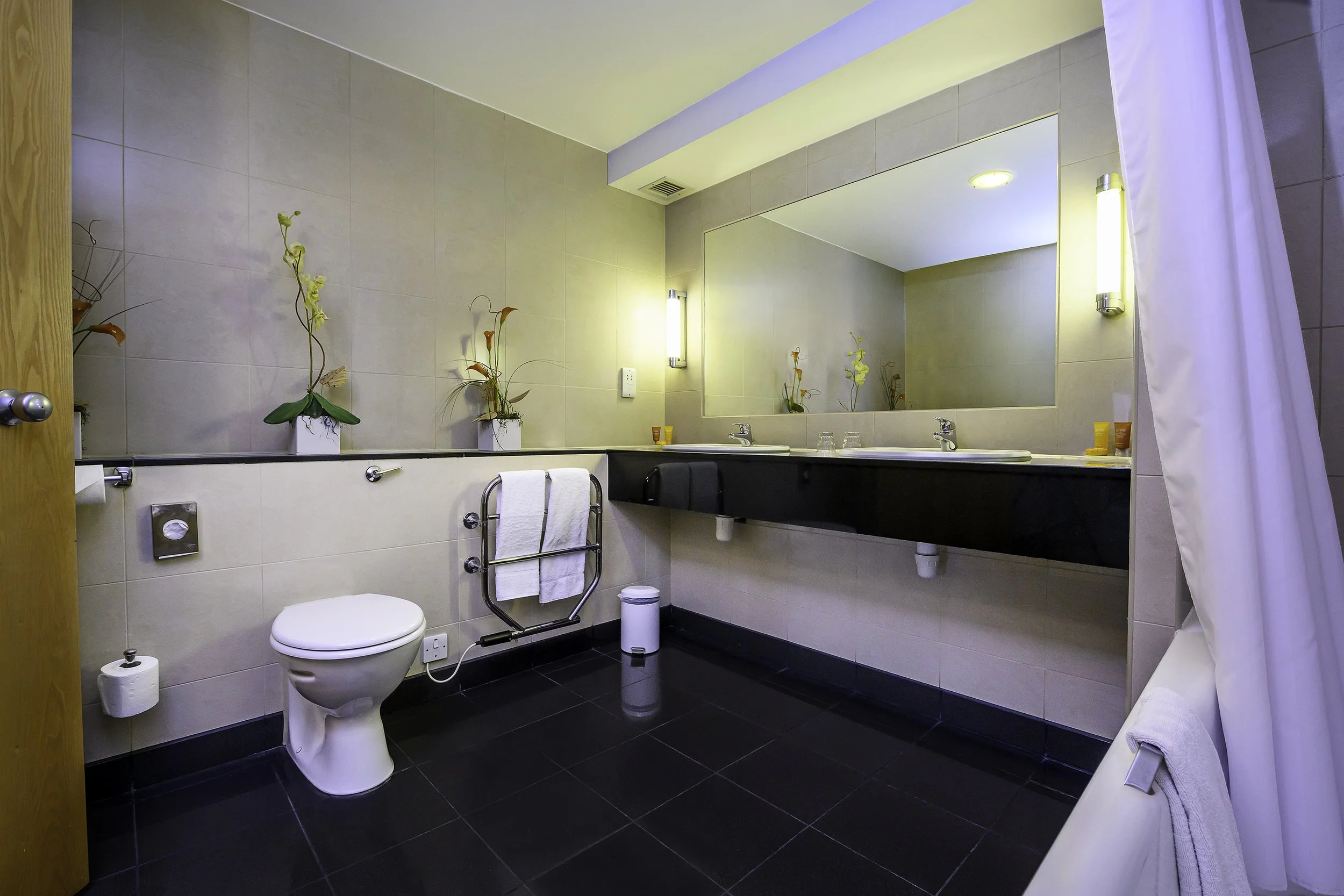 A clean bathroom with beige tiled walls, a large mirror above two sinks, and black countertop. There is a white toilet, a towel hanging on a metal rack, and decorative plants on the counter and shelf. Soft lighting and a shower curtain are visible.