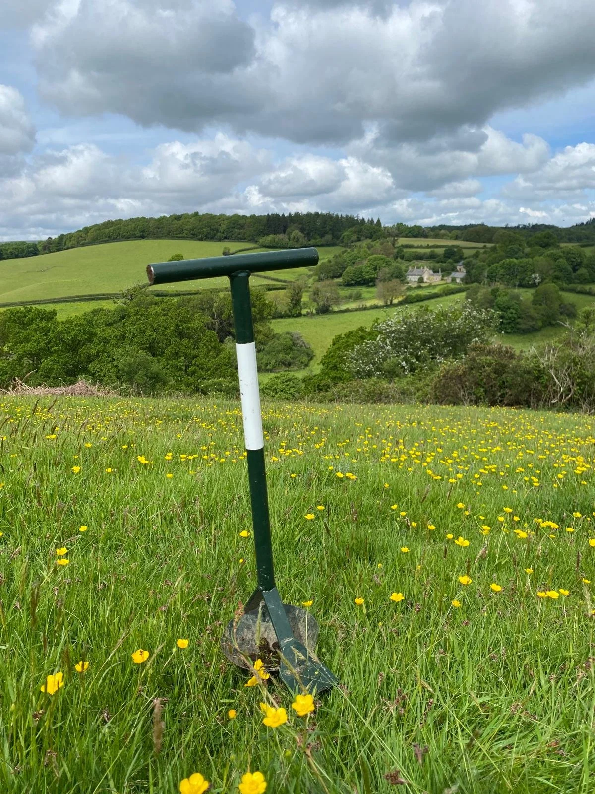 A metal digging tool stands upright in a grassy field filled with yellow flowers, with a landscape of rolling green hills, trees, and some houses under a partly cloudy sky in the background.