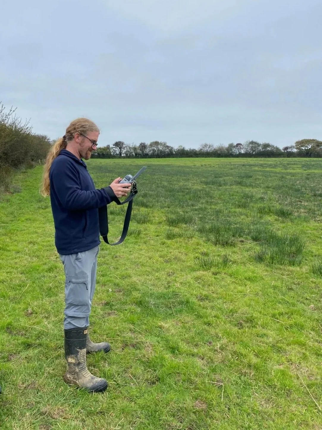 Man standing in a grassy field, holding a remote controller, wearing rubber boots, glasses, navy hoodie, and light-colored pants, with trees and cloudy sky in the background. Drone survey and design.