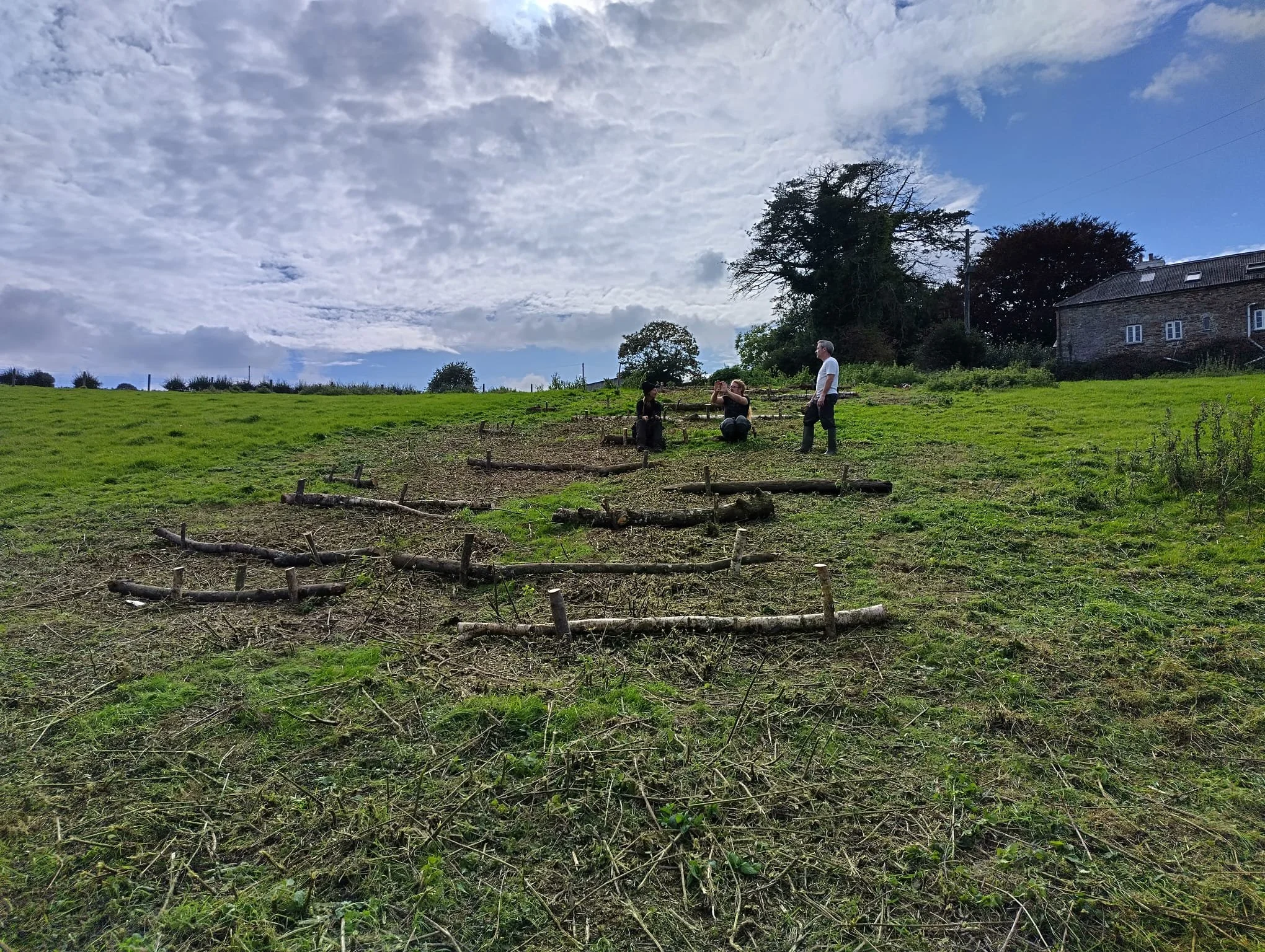 A grassy hillside with a few people, some standing and some sitting, under a partly cloudy sky. There are trees and a house in the background. conservation services South West England, environmental consultancy South West, practical installations