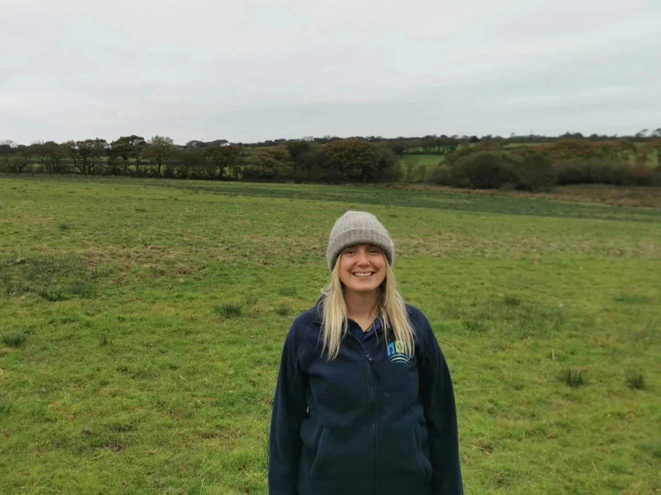 A young woman with long blonde hair, smiling, wearing a gray knit hat and a navy blue jacket, standing in a green open field with some trees and hills in the background under a cloudy sky. Ecological surveys