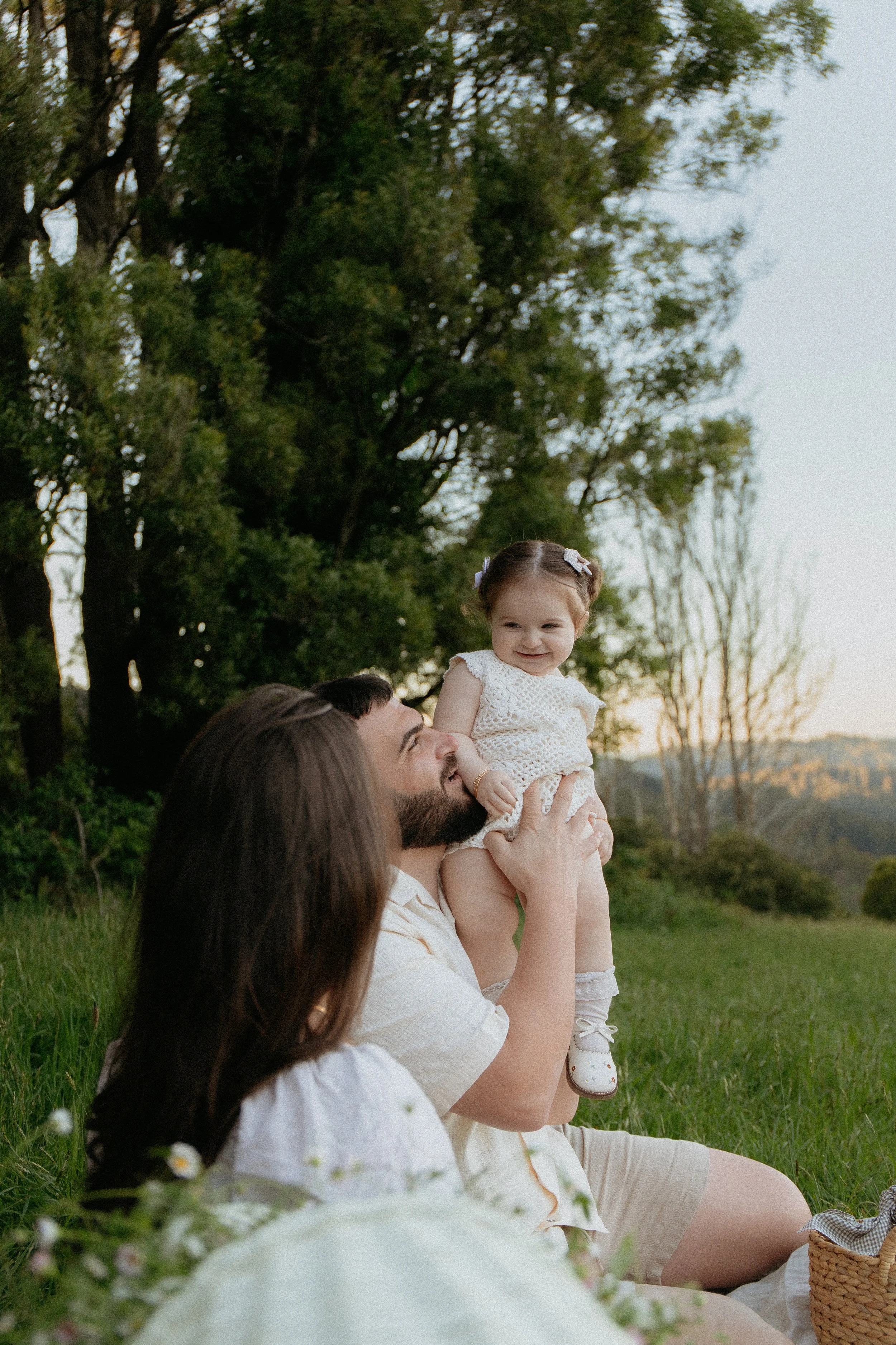 a family of three in surrounded by greenery, smiling and dad holding daughter up