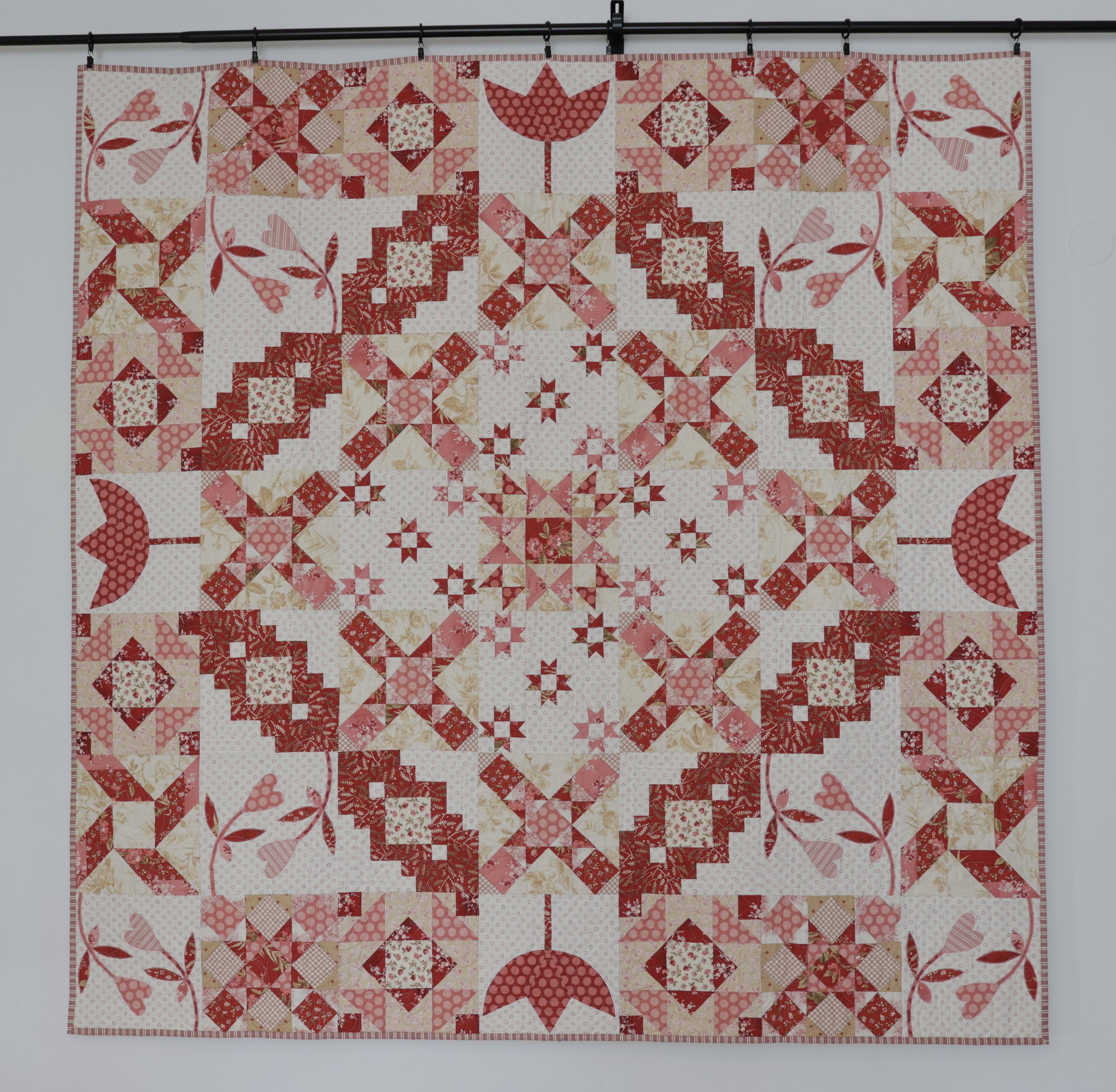 A large quilt with red, pink, cream, and beige patches arranged in a symmetrical, intricate pattern featuring stars, hearts, flowers, and animals, hanging from a black rod on a white wall.