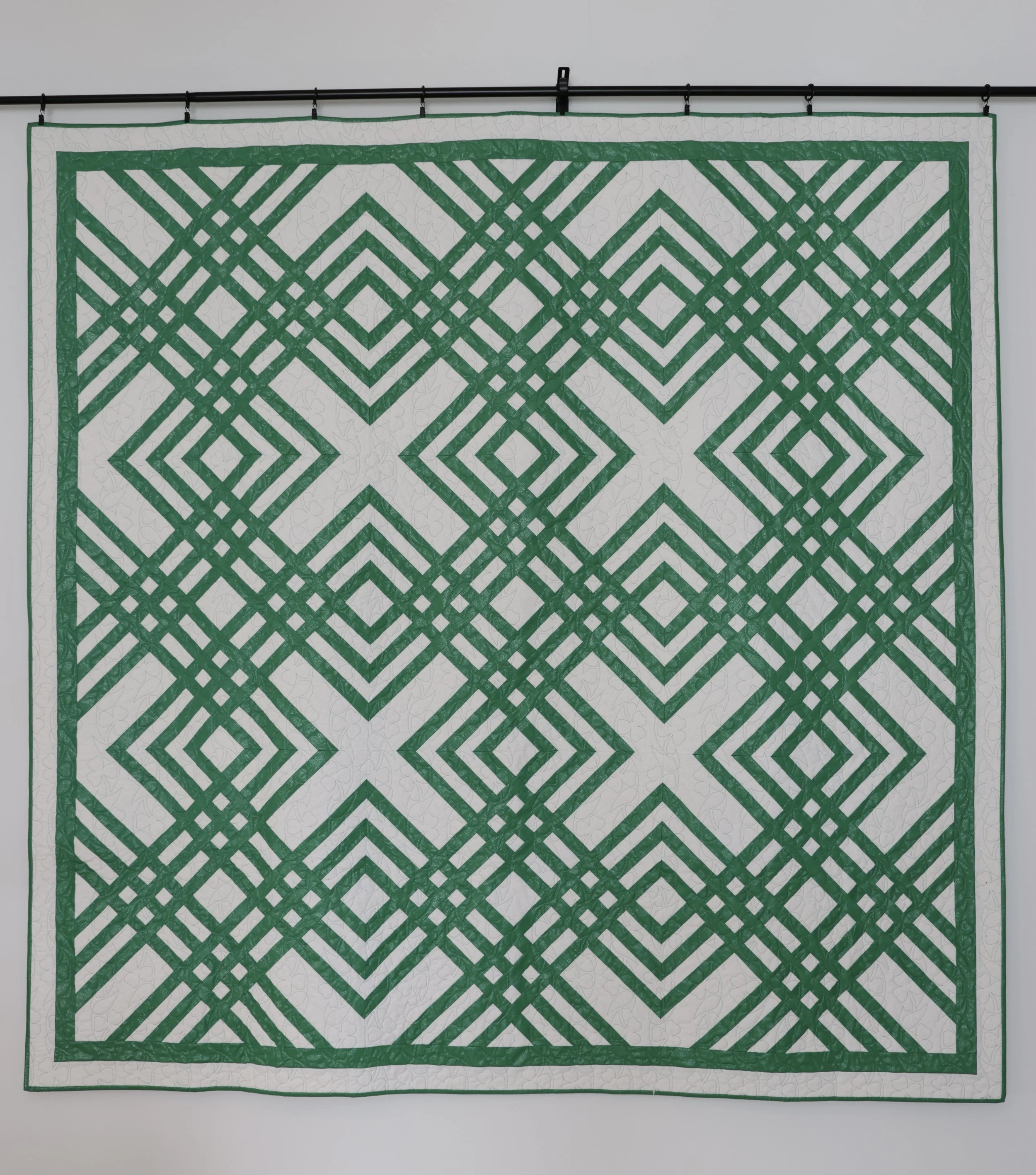 A quilt with a geometric green pattern on a white background, hanging from a black rod against a plain wall.