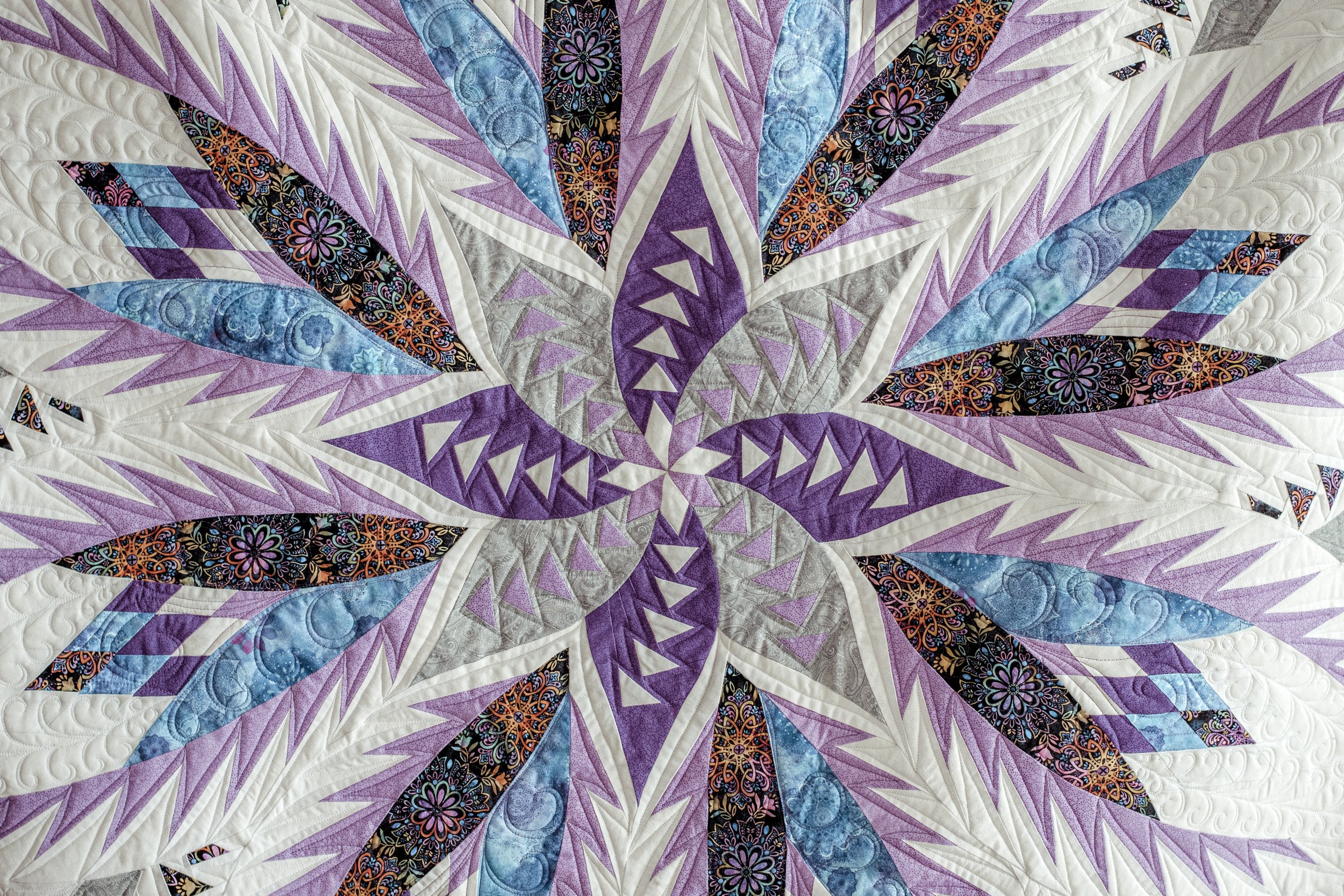 A colorful quilt featuring a floral, starburst pattern in shades of purple, blue, black with floral designs, white, and gray fabric.